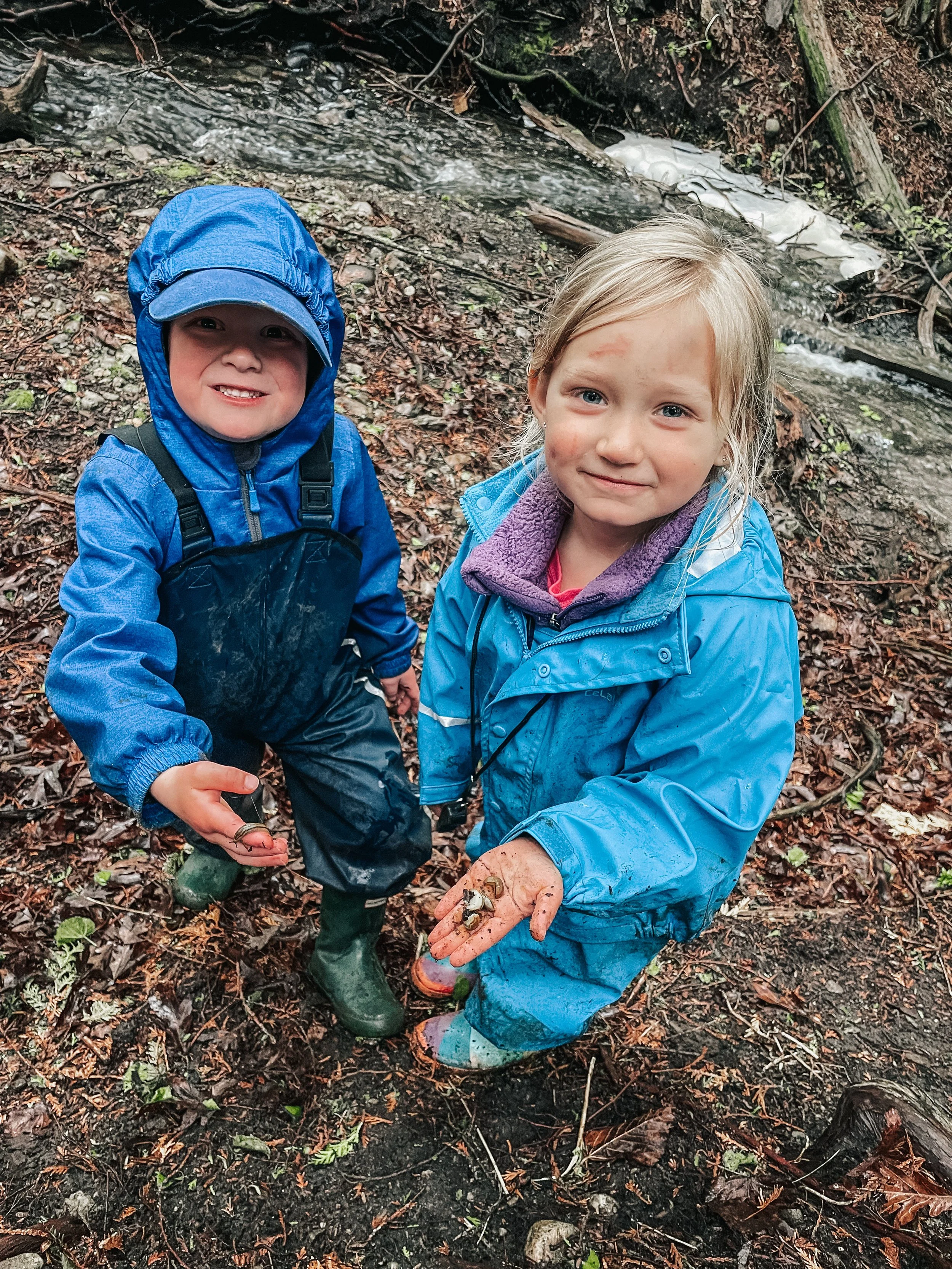 Two children, a boy and a girl, standing outdoors in a muddy, wooded area near a small creek, holding small frogs in their hands and smiling.