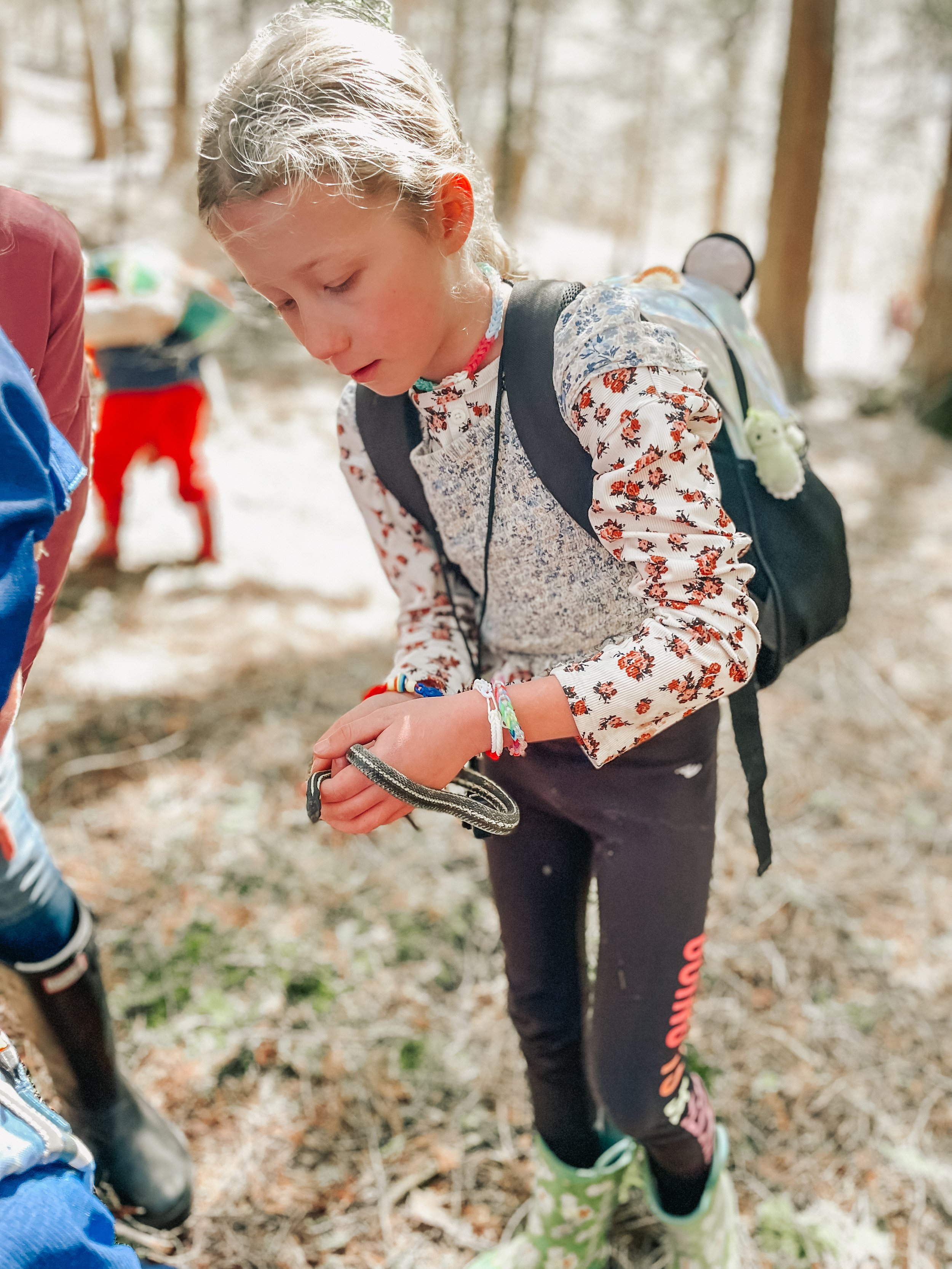 A young girl with blonde hair in a ponytail, wearing a floral long-sleeve shirt, black pants, and green rain boots, holding a snake in a forest.
