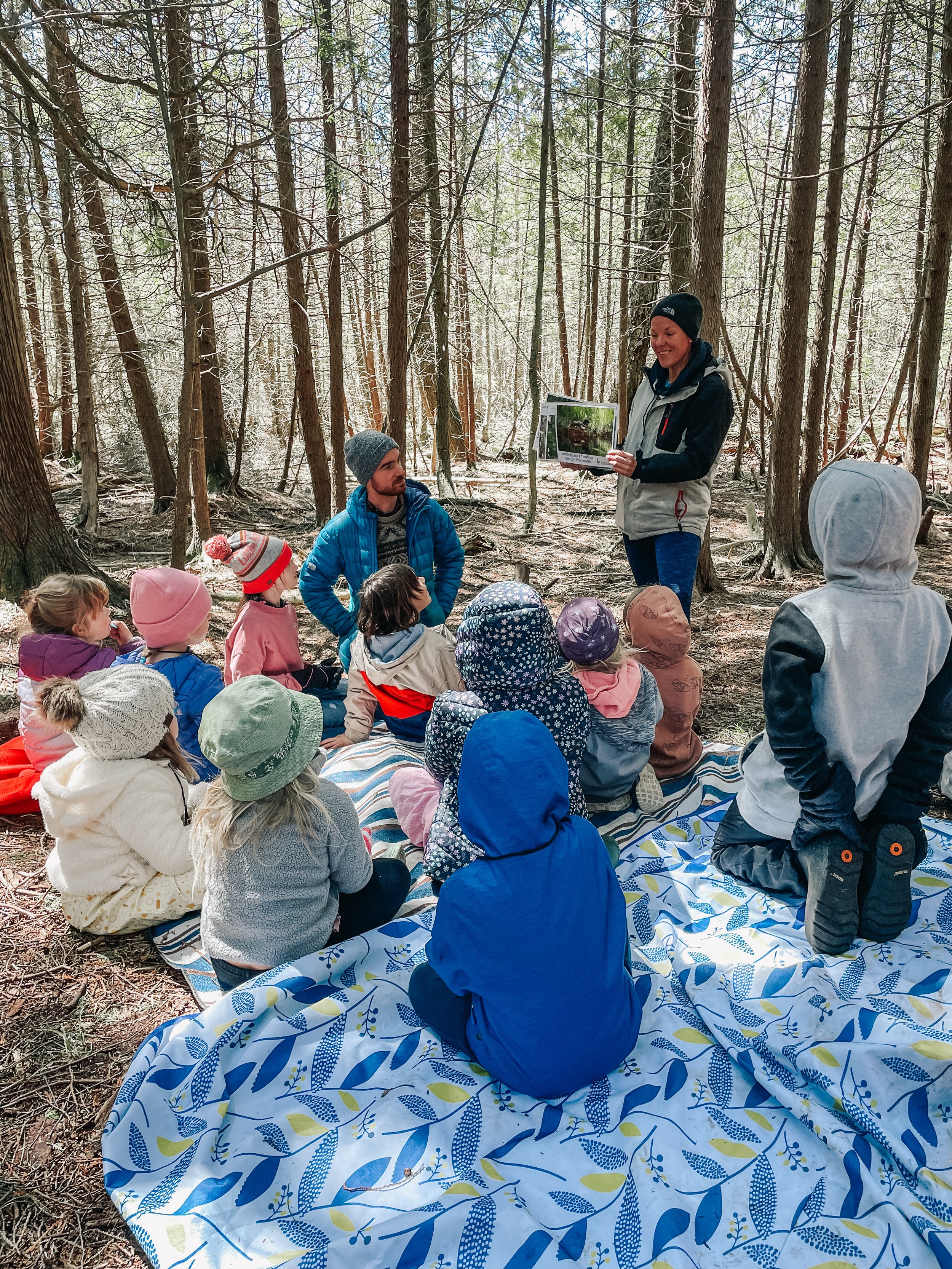 Group of children sitting on a blanket in a forest, listening to a woman holding a picture, while a man looks on.