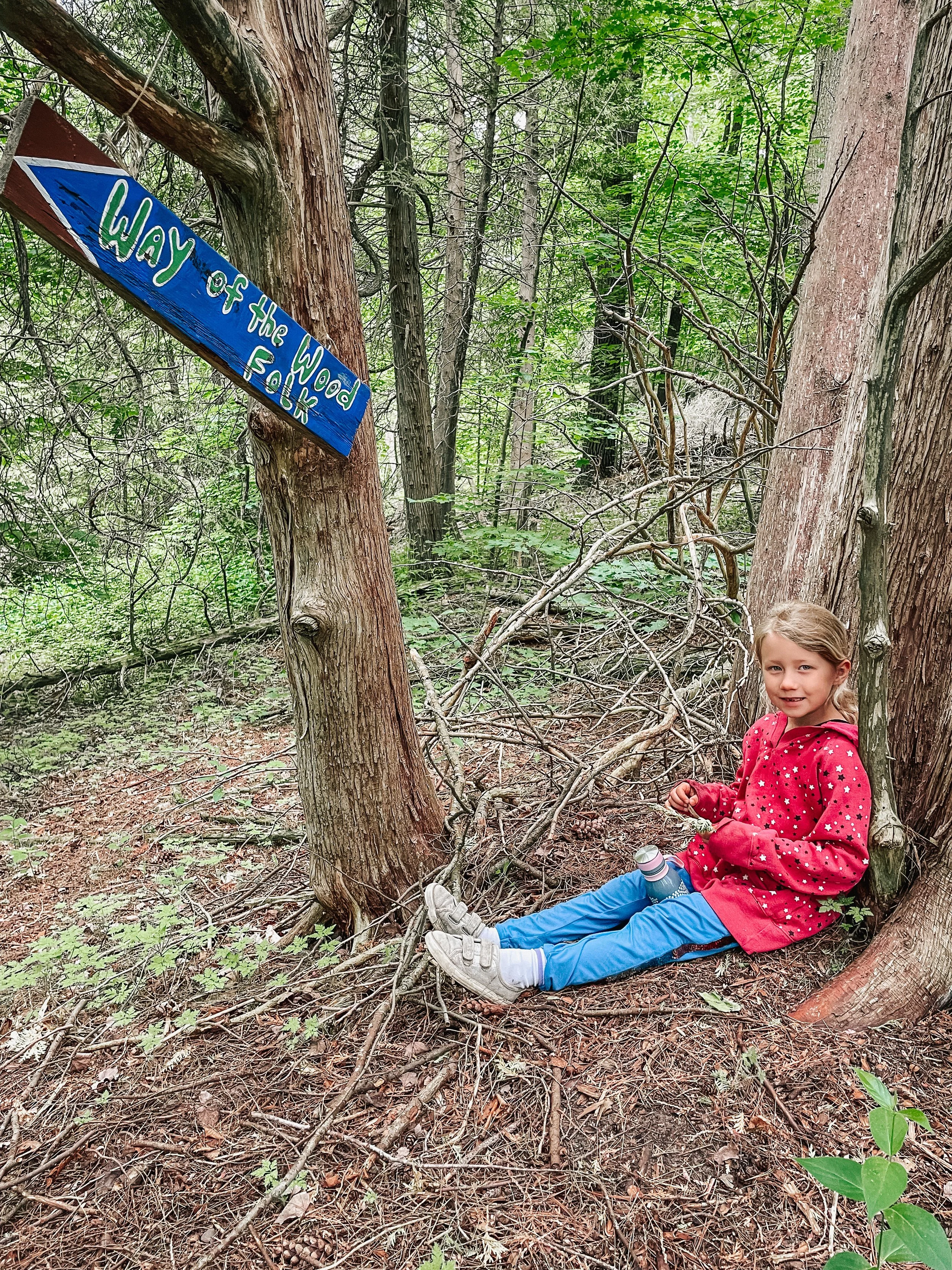 A young girl sitting against a tree in a forest, holding a water bottle and smiling. A sign on a tree reads 'Way of the Wood Fork'.
