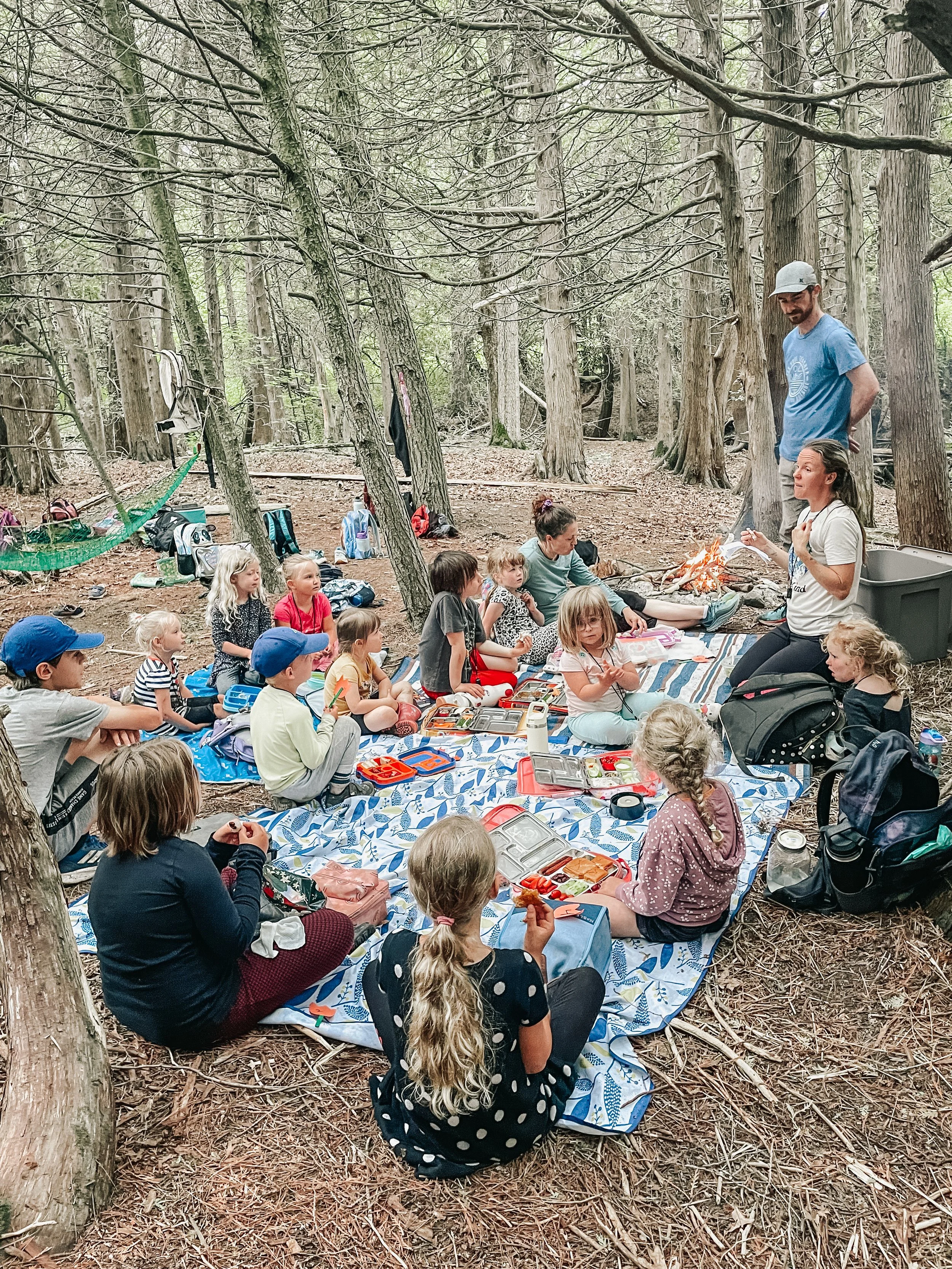 Group of children and adults having a picnic in a wooded forest, sitting on a large blue and white patterned blanket surrounded by trees, backpacks, and lunch containers.