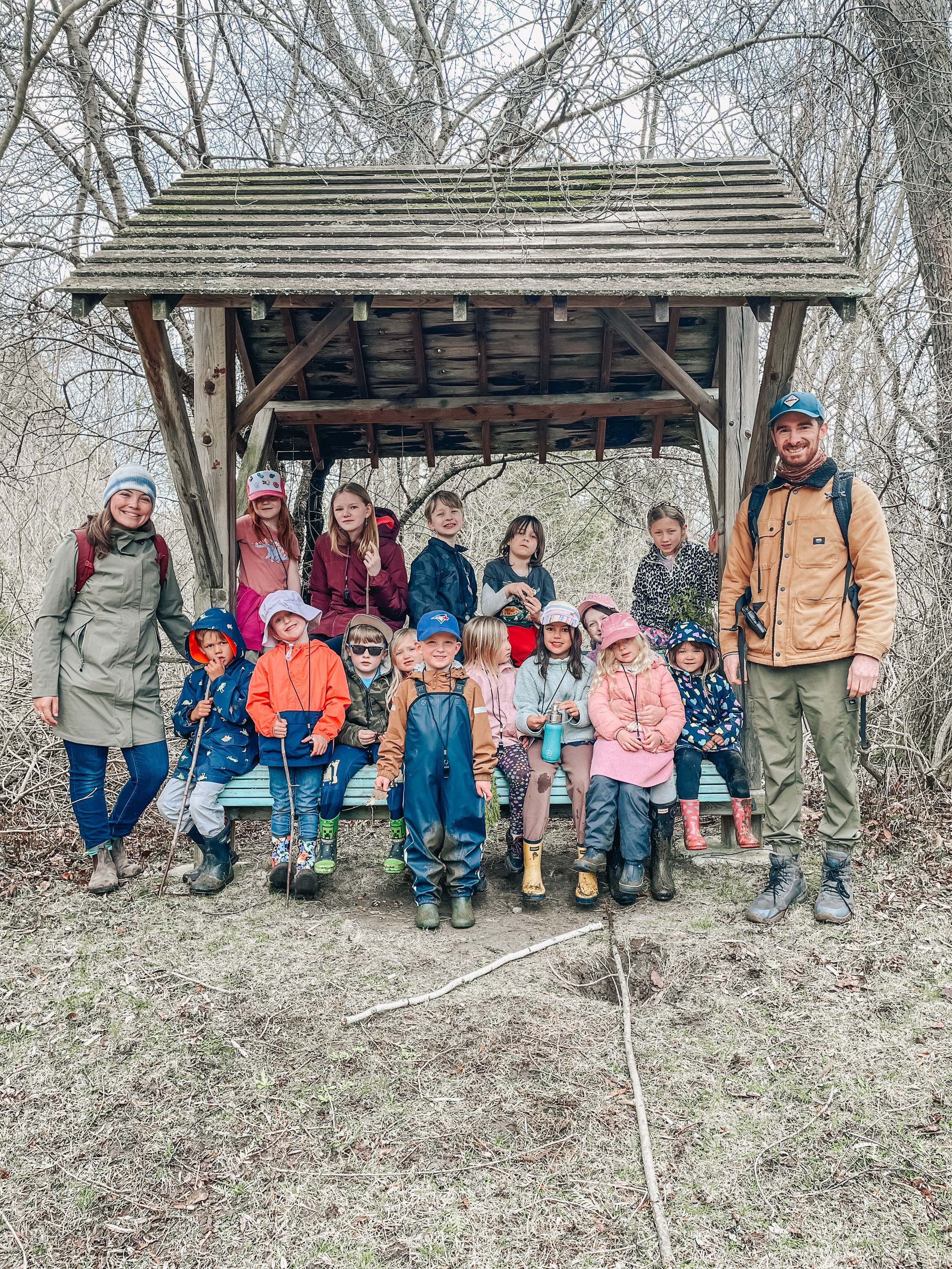 A group of children and two adults posing together under a wooden shelter in a wooded area during a hike or outdoor activity.