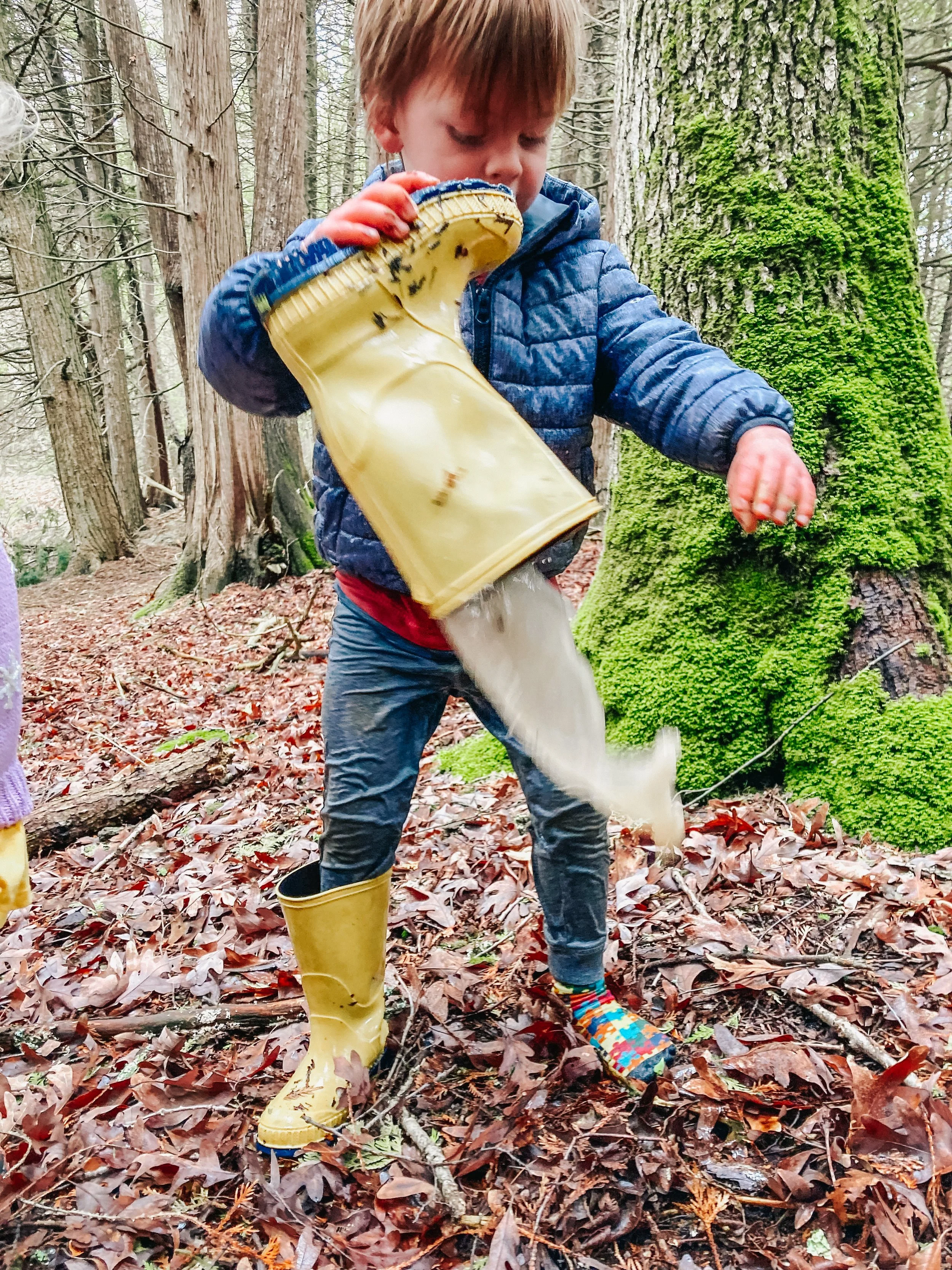 Child wearing yellow rain boots and a blue jacket playing in a forest and pouring water from a yellow watering can.