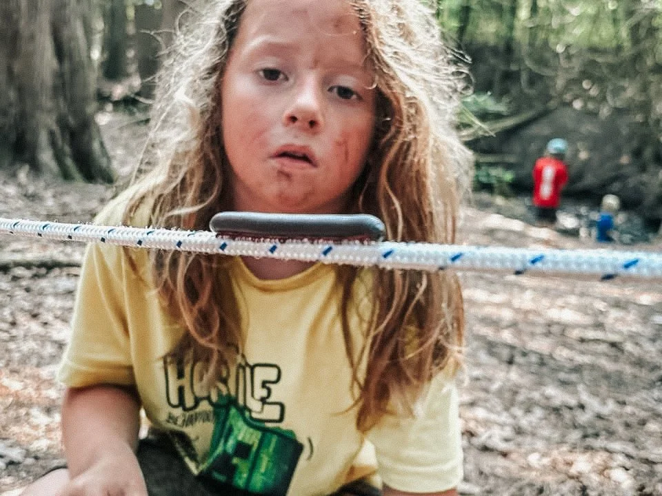 A young girl with long red hair and a yellow shirt looks at a smartphone balanced on a rope in a forest.