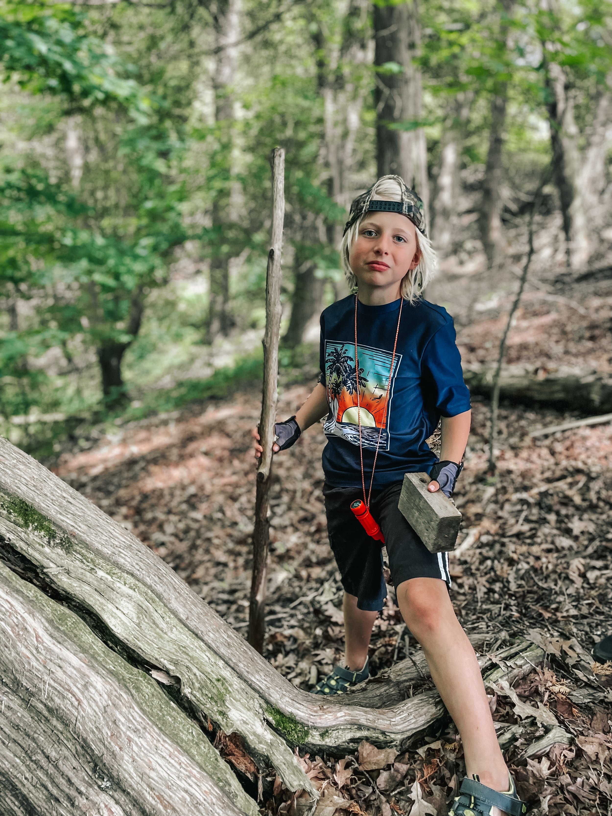 A young boy with long blond hair, wearing a backwards cap, navy blue t-shirt with a sunset and palm trees graphic, black shorts, and sandals, stands outdoors in a wooded area holding a walking stick and a rock. He appears to be exploring or playing i