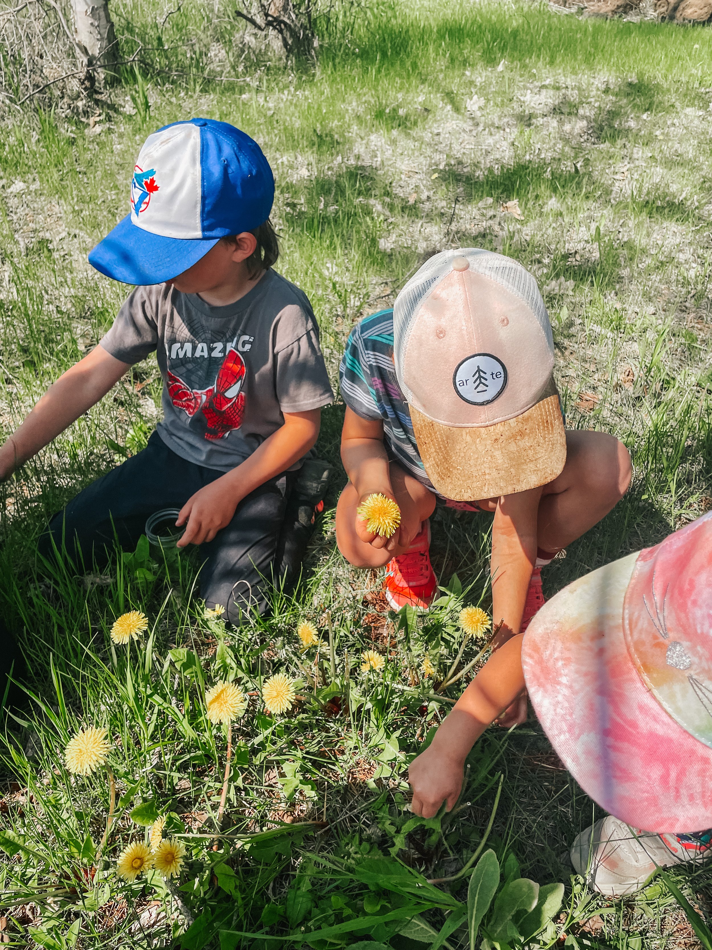 Three children exploring a grassy field with yellow flowers, wearing caps and casual clothing.