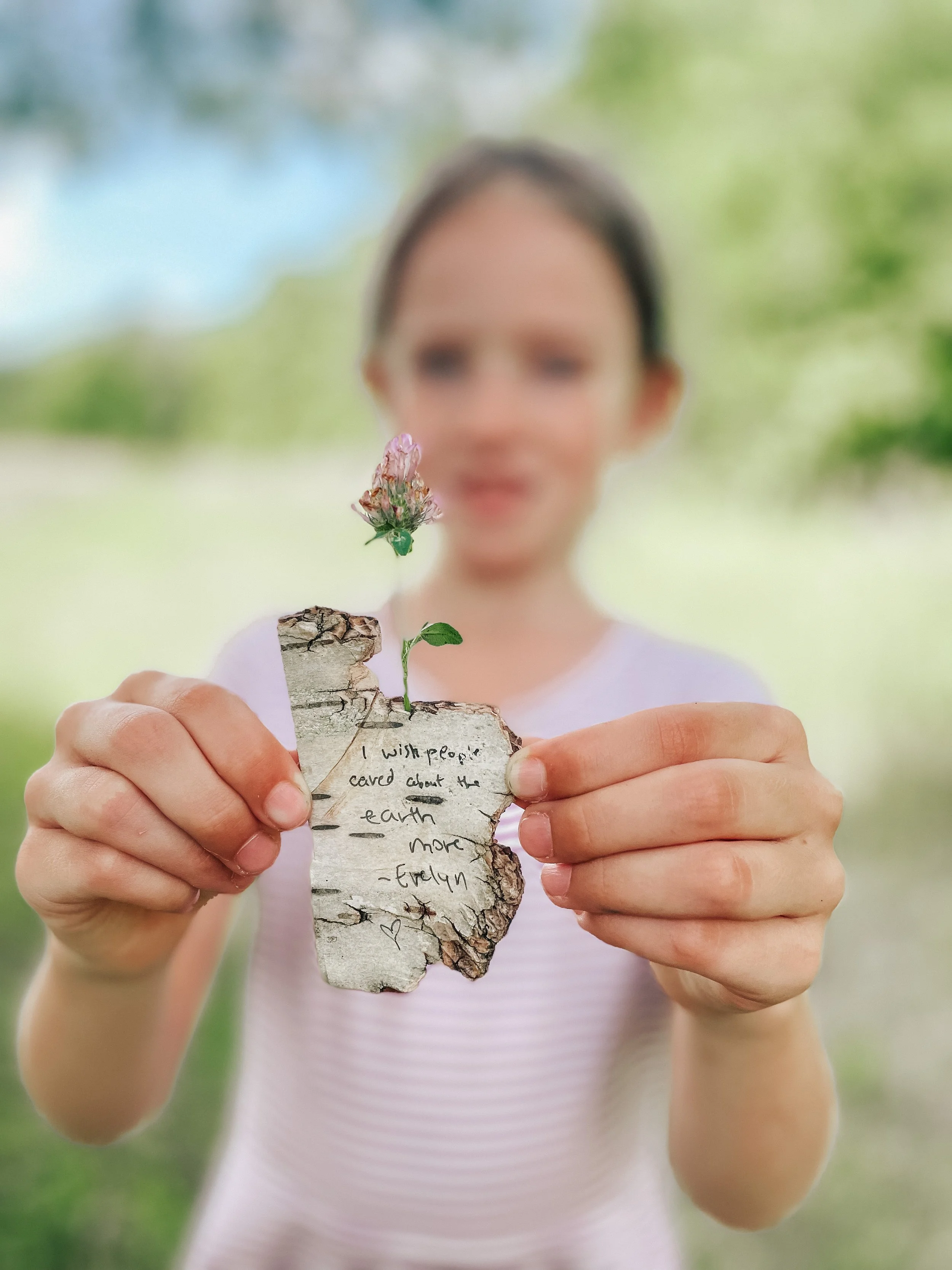 A person holding a piece of bark with a small sprout and a pink flower growing from it. The bark has a handwritten note that says, 'I wish people cared about the earth more - Evelyn'.
