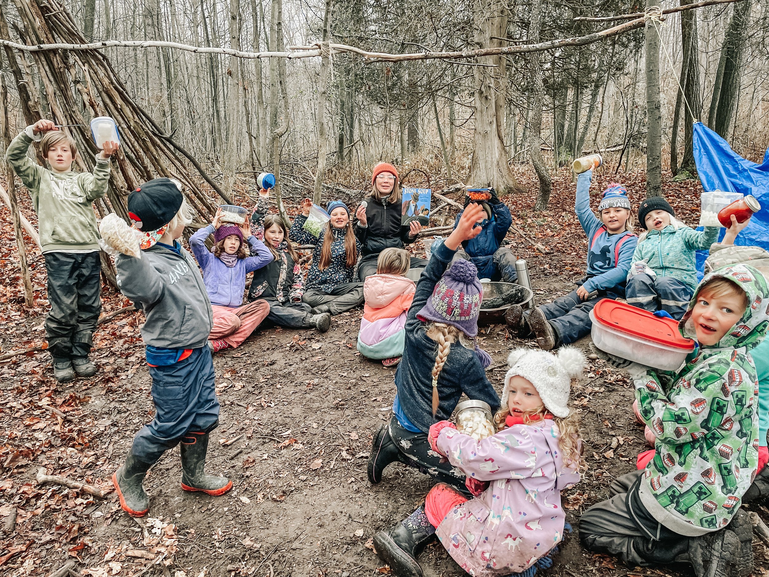 Group of children and a woman sitting in a forest, surrounded by trees, holding containers of food or snacks, with a makeshift shelter made of branches behind them, during a fall or winter day.