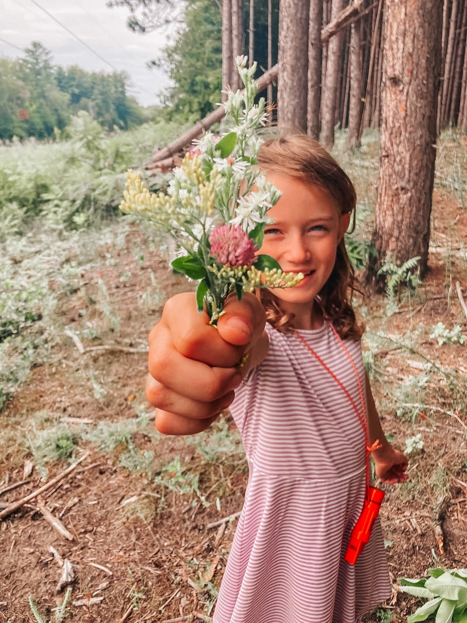 A girl with long hair wearing a striped dress holding out a bouquet of wildflowers in a forest.