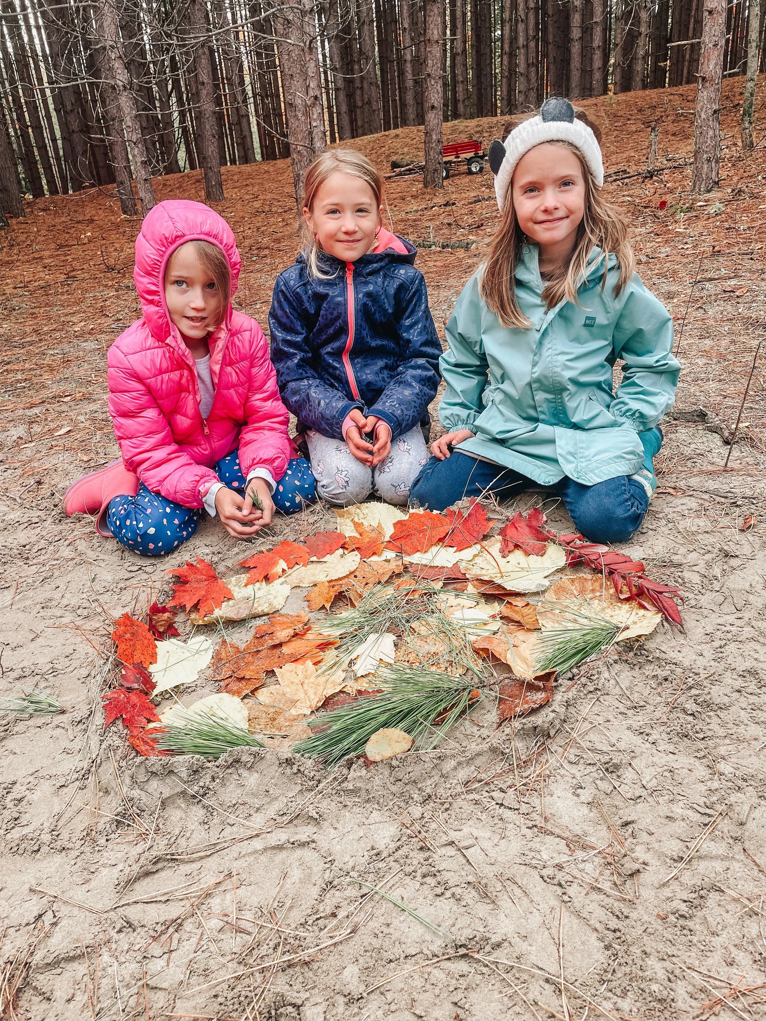 Three young girls kneeling on the ground in a wooded area with autumn leaves and pine branches arranged in a circle in front of them, wearing jackets and hats.