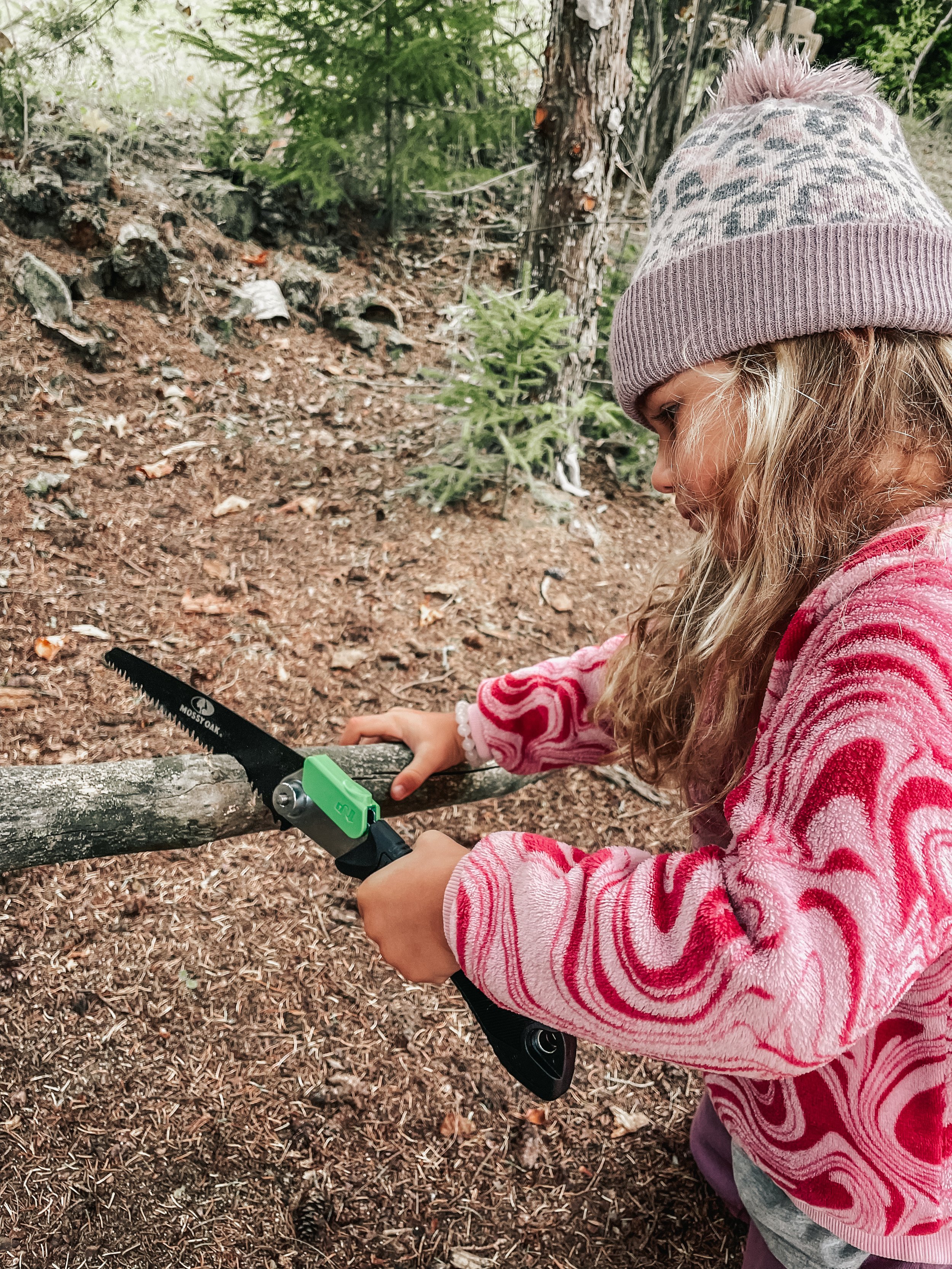 A young girl wearing a pink patterned jacket and a knit hat is outdoors in a wooded area, cutting a small tree branch with a saw.
