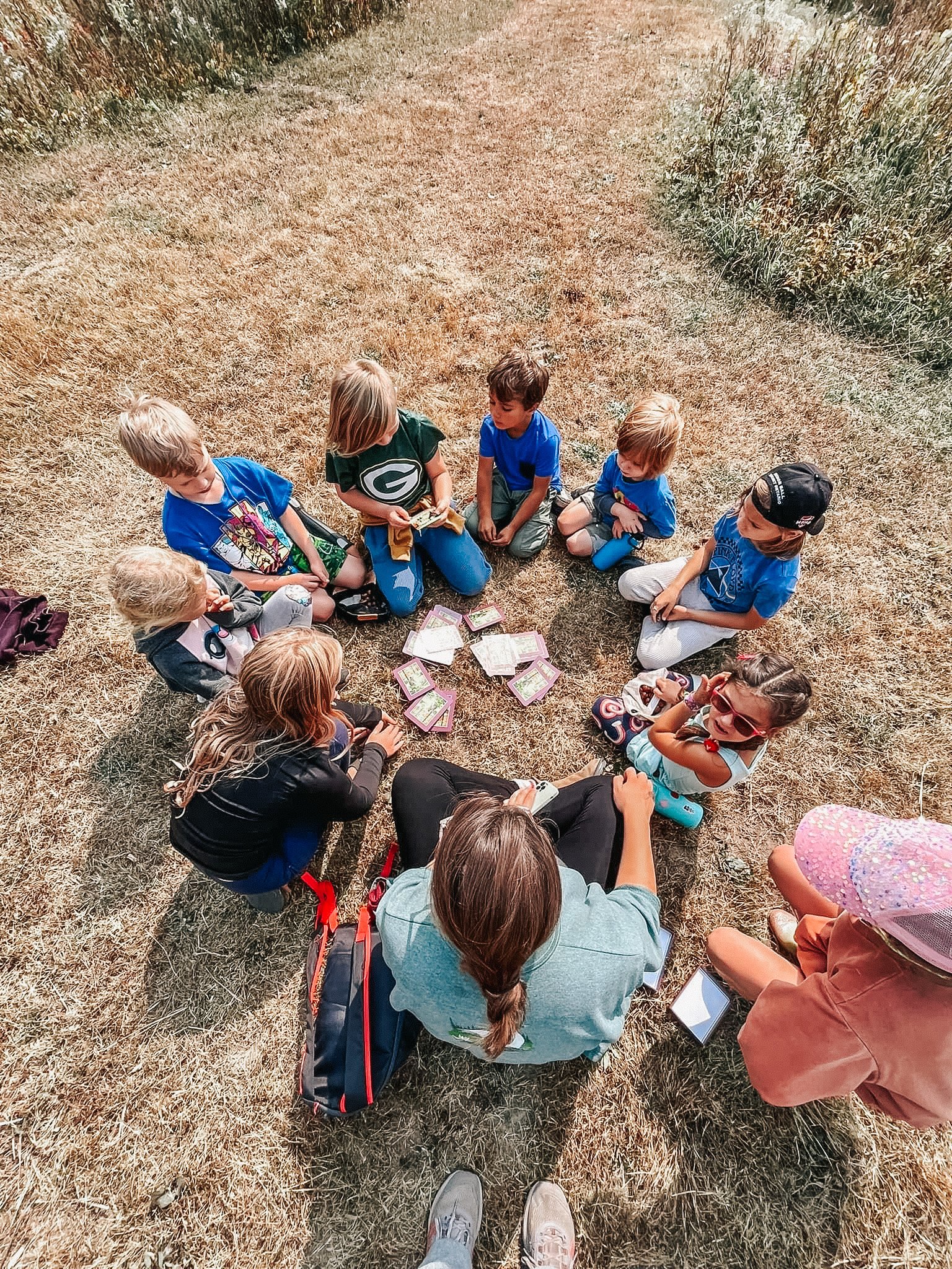 A group of children and a woman sitting in a circle on a dry grassy field, playing a card game outdoors in daylight.