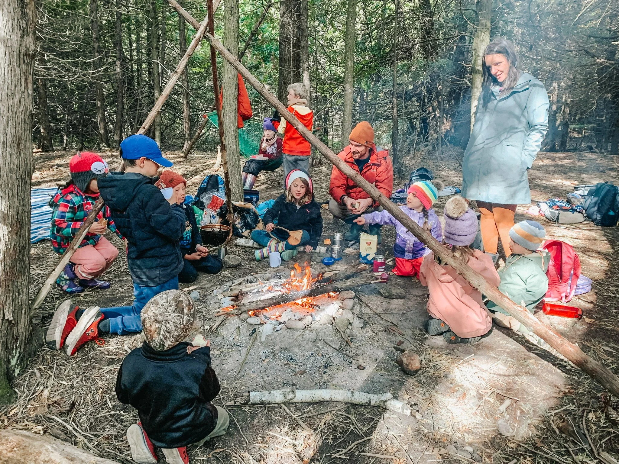 Children and adults gathered around a campfire in a wooded area, some sitting on the ground and some standing, with backpacks and outdoor gear around them.