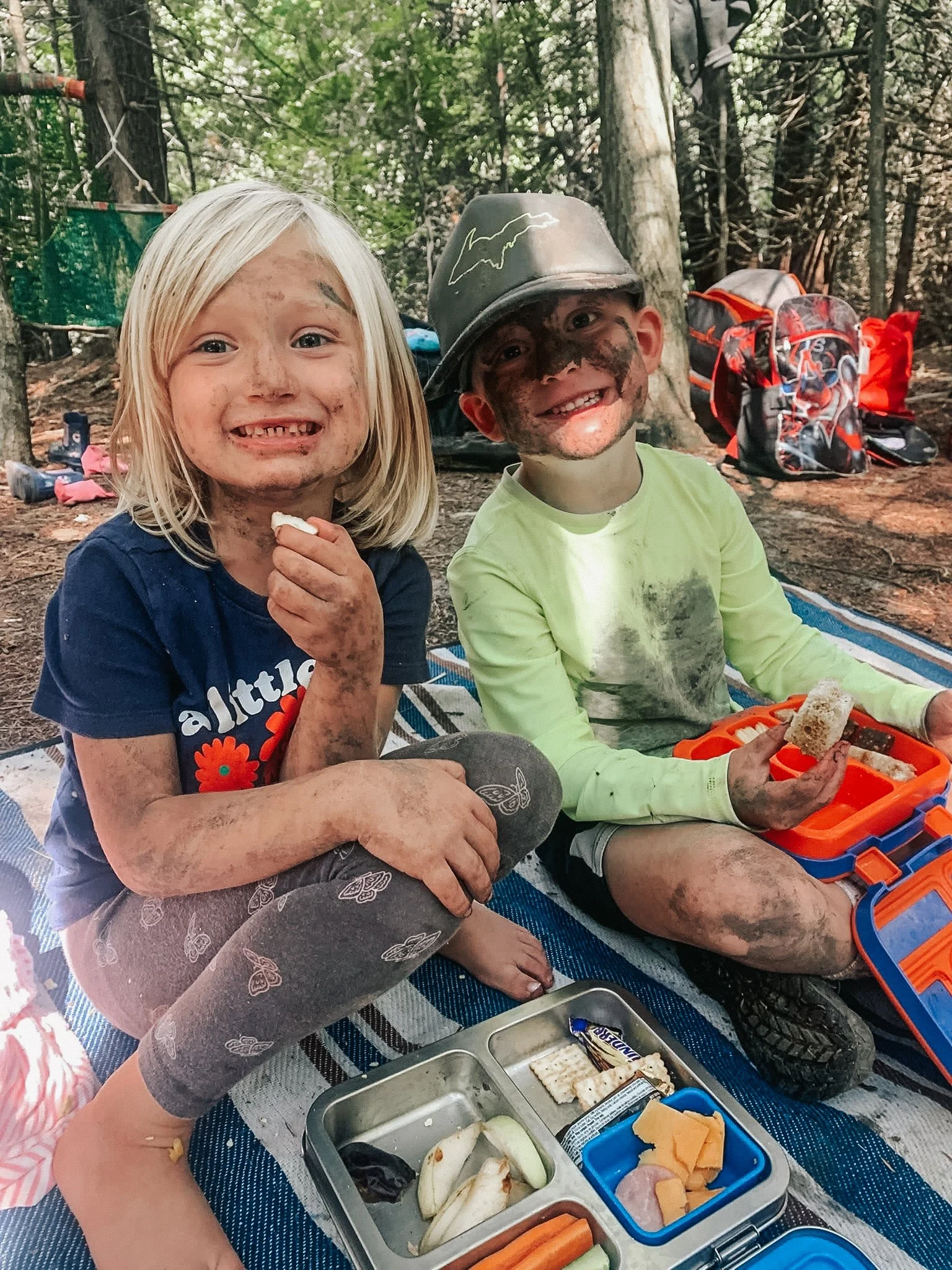 Two children, a girl and a boy, sitting on a picnic blanket in the woods, covered in dirt and mud, smiling and enjoying a snack of bread and cheese with a picnic lunch box in front of them.