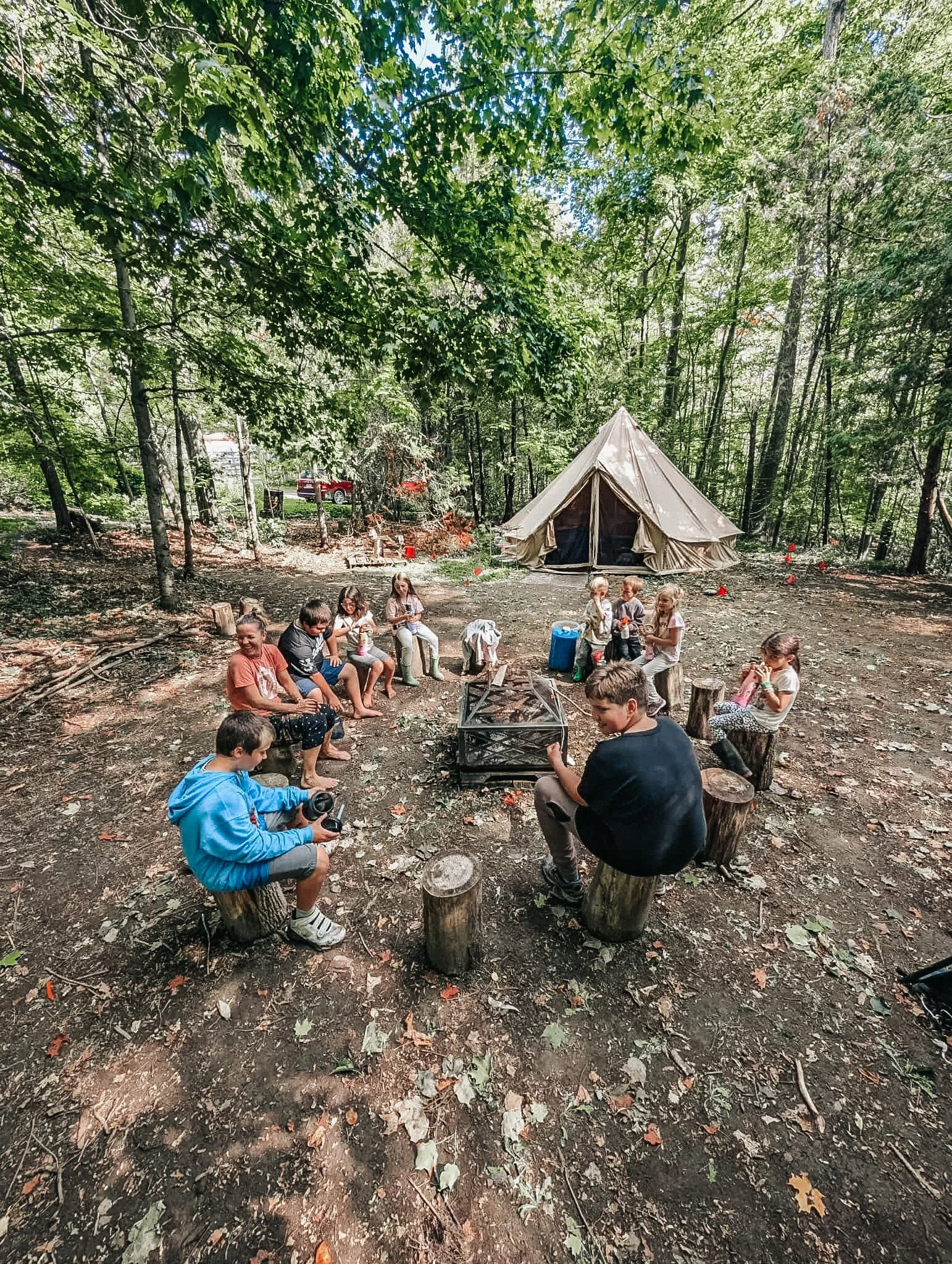 Children sitting around a campfire in a wooded outdoor setting, with a canvas tent in the background.
