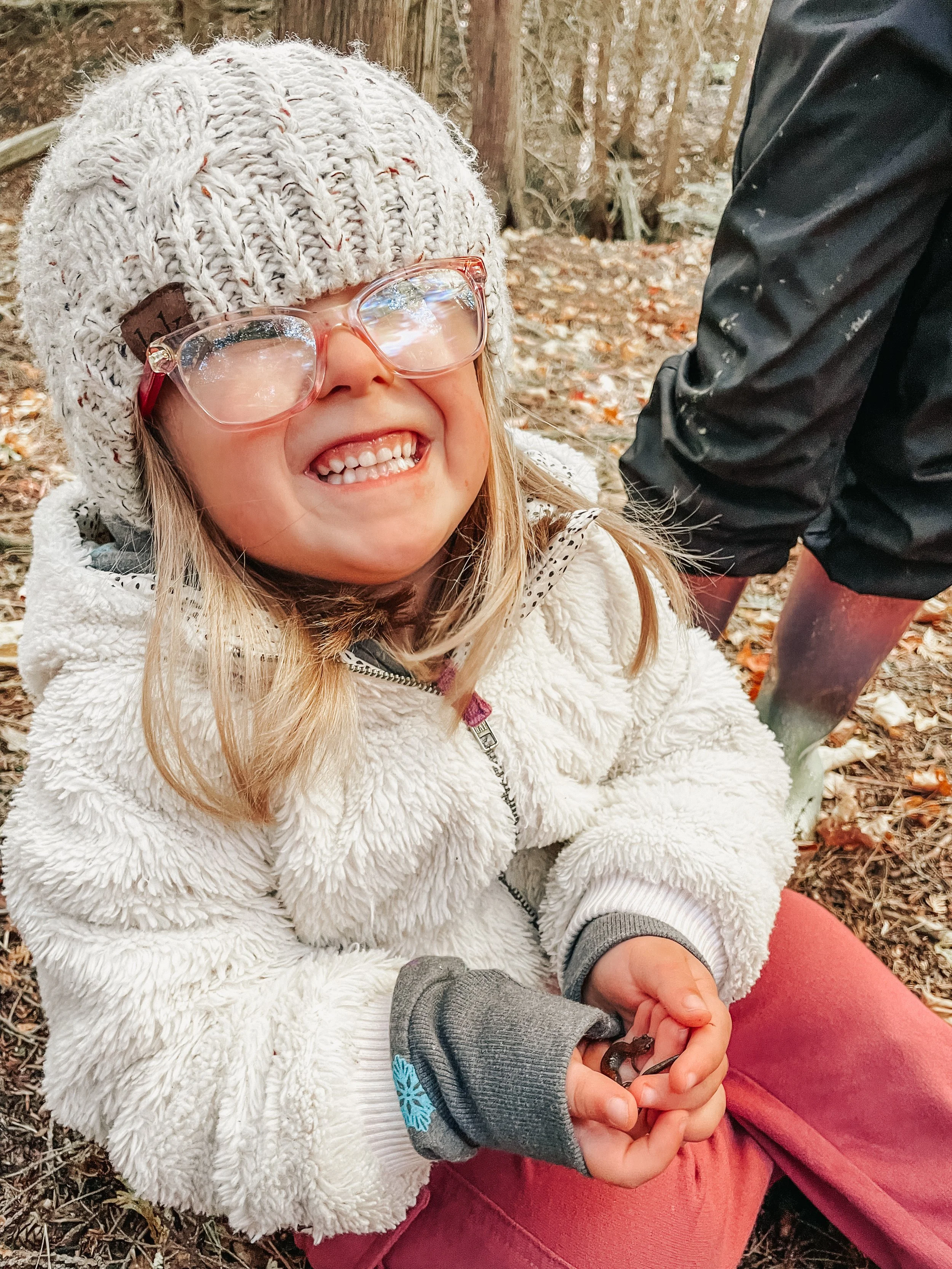 A young girl smiling happily while sitting outdoors in a wooded area, wearing glasses, a knitted beanie, a white fuzzy jacket, and reddish pants. She's holding a small object in her hands.