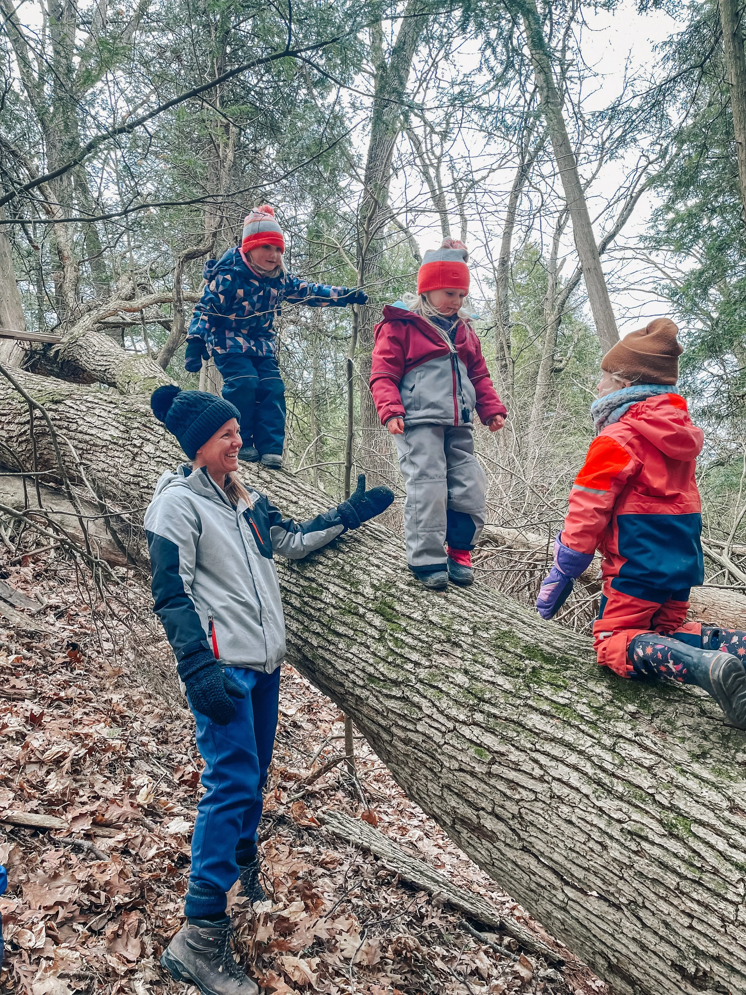 Three children and a woman in winter clothing play on a fallen tree in a wooded area during the daytime, with two children standing on the tree and one sitting on it, while the woman supports and interacts with the children.