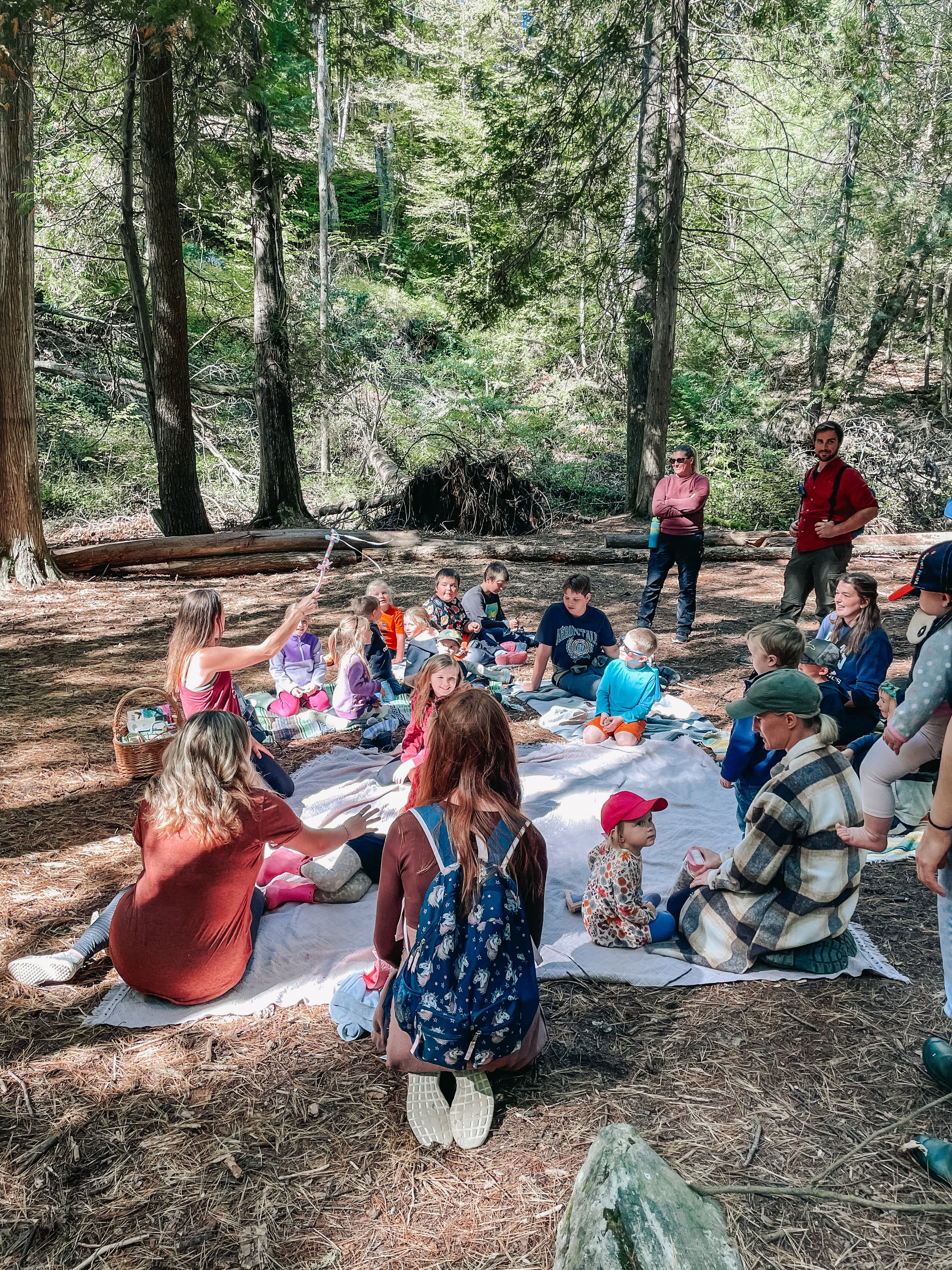 A group of children and adults sitting on blankets in a forest clearing, participating in an outdoor activity or storytelling session, surrounded by tall trees and sunlight filtering through the leaves.