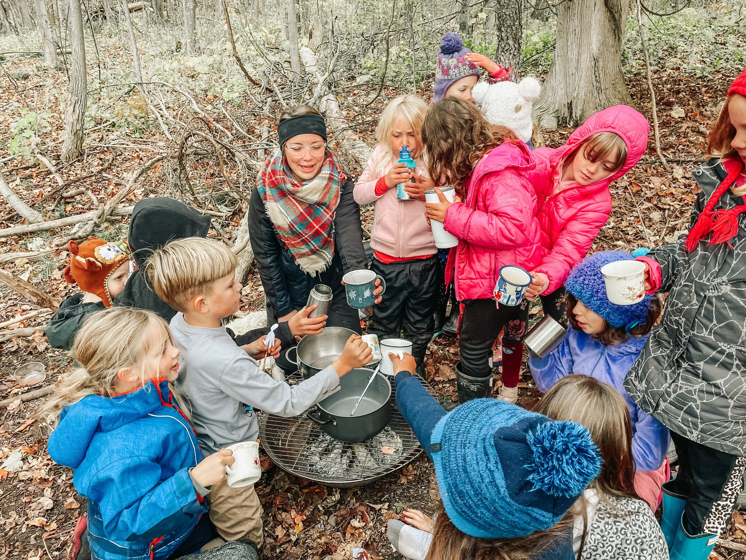 Children and adults outdoors in a forest, gathered around a campfire cooking and drinking hot beverages, dressed in warm clothing and hats.