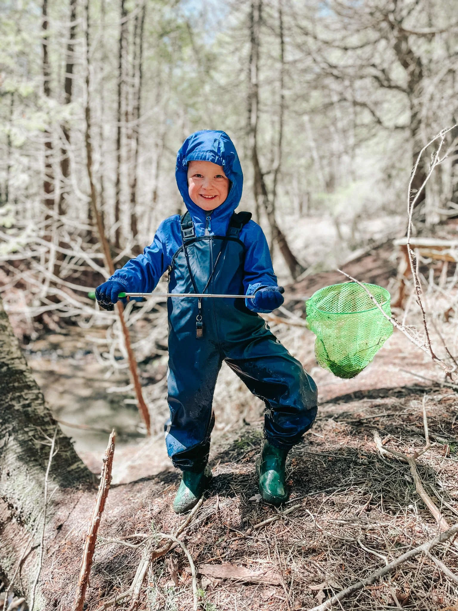 A young boy wearing a blue raincoat, rain boots, and gloves, holding a green net, standing in a wooded area during a nature exploration.