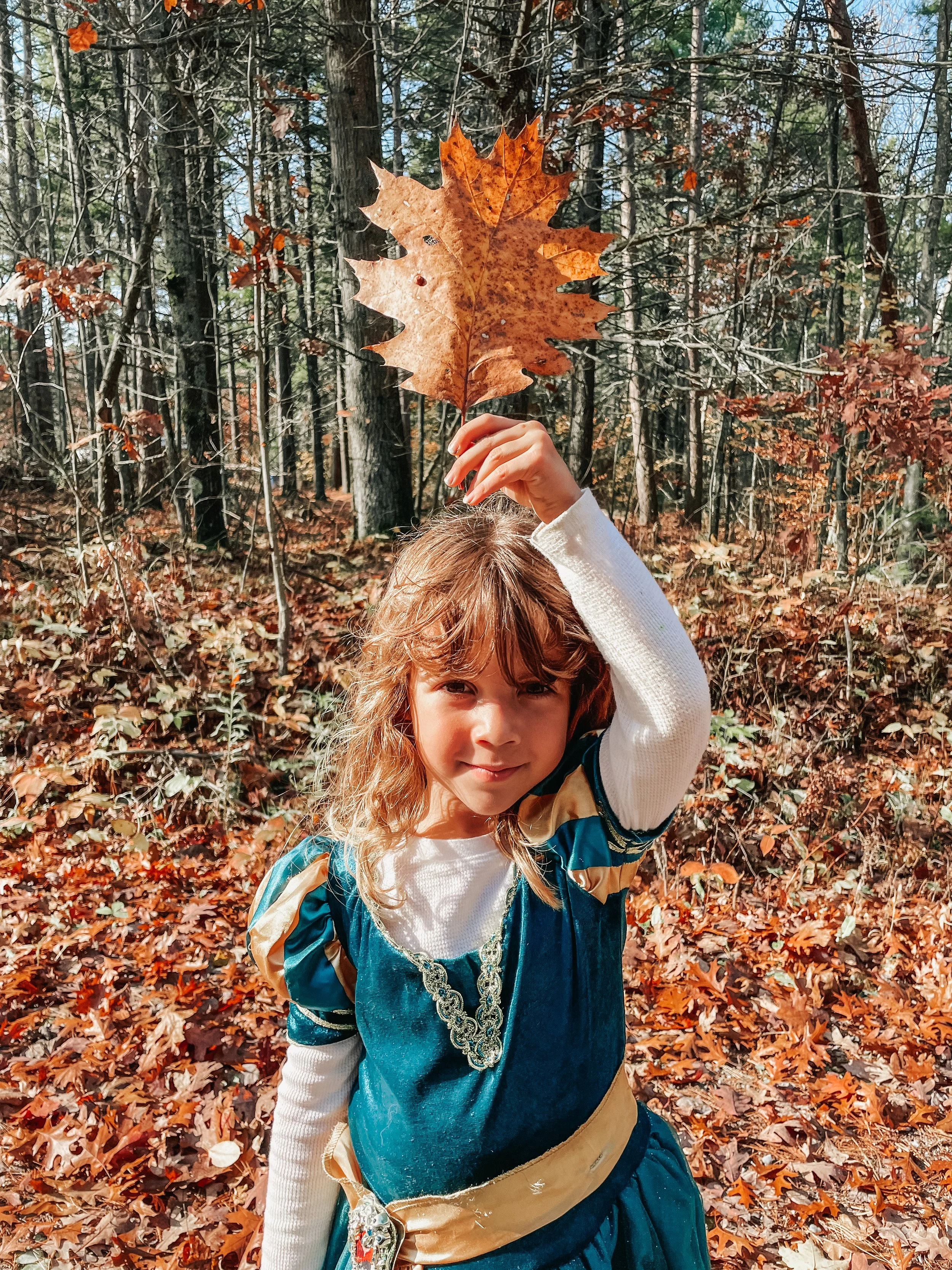A young girl with curly hair in a blue dress with gold accents, holding a large brown maple leaf above her head in a forest during autumn, with fallen leaves on the ground.