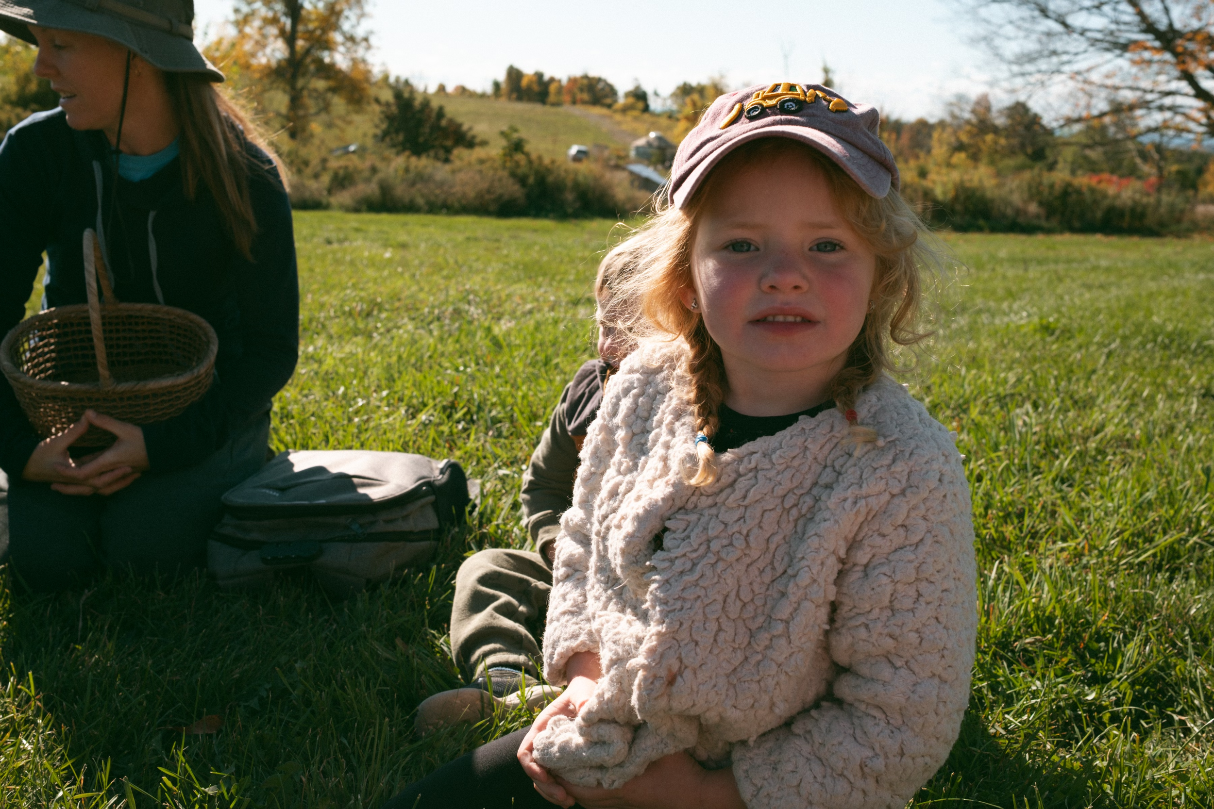 A young girl with blonde curly hair sitting on the grass outdoors, wearing a pink cap with a truck design and a fluffy beige jacket, looking at the camera with a slightly confused or concerned expression. In the background, there is a woman with a hat holding a basket, and a backpack on the ground. The setting is a grassy field with trees and a hill in the distance under clear sky.