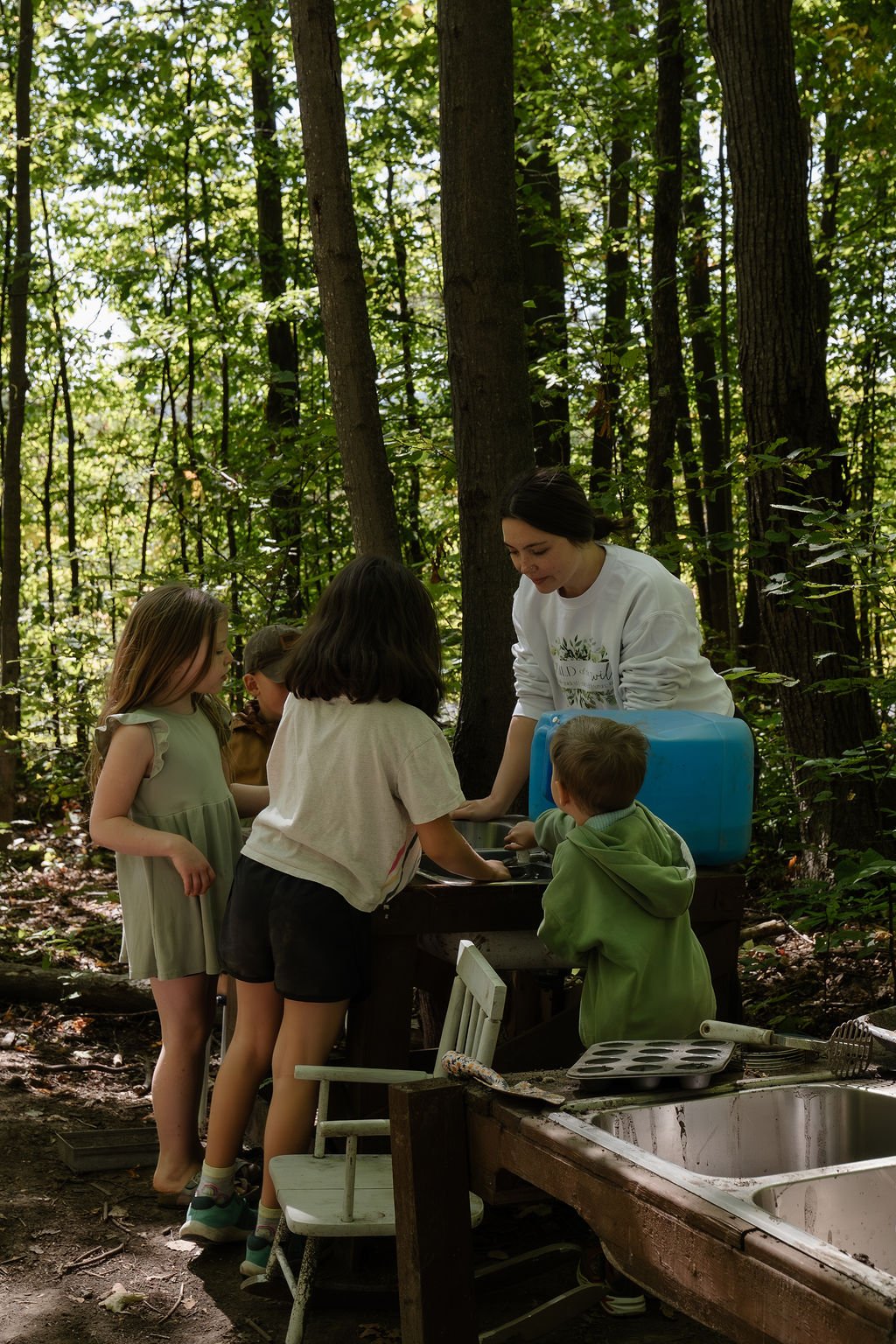 A group of children and a woman gather around an outdoor sink station in a wooded area, engaged in a hands-on activity, possibly cooking or washing items.