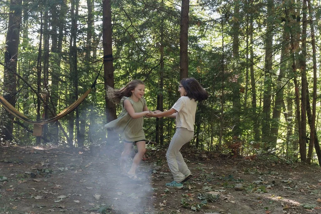 Two young girls playing and dancing in a forest, holding hands, with sunlight filtering through the trees, and a hammock hanging in the background.