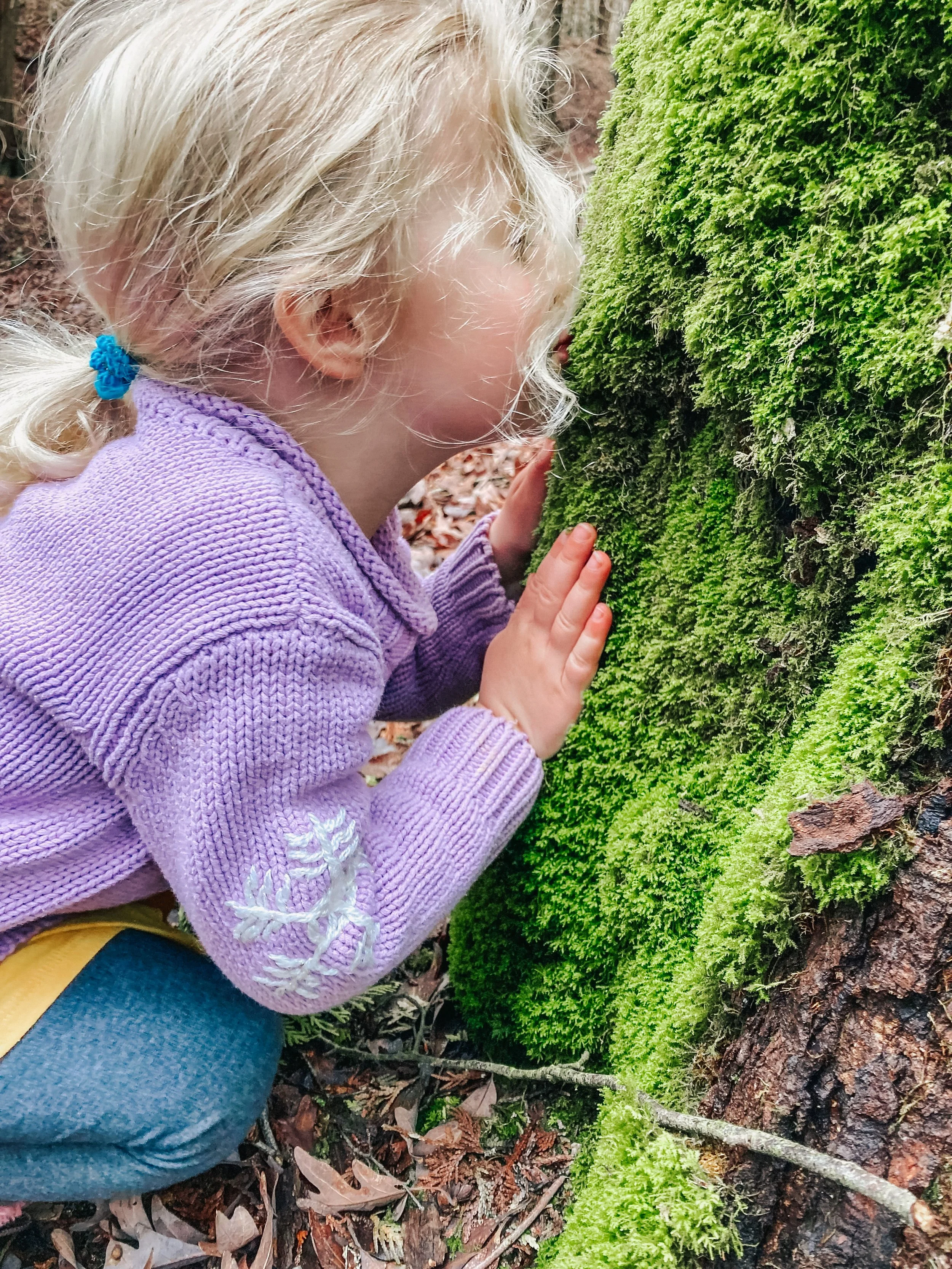 A young girl with blond hair, wearing a purple sweater with white embroidery on the sleeve, is crouching and touching bright green moss on a tree trunk in a forest.