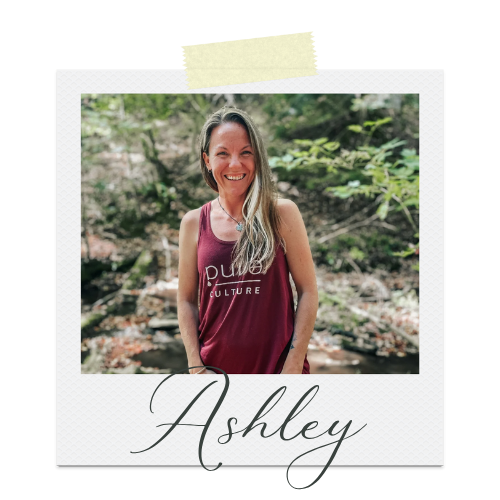 A woman with long blonde hair smiling outdoors, wearing a sleeveless maroon top with the words 'Pure Culture' printed on it. The background features trees and a natural setting. The photo is framed with a white polaroid-style border with the name 'Ashley' written in cursive at the bottom.