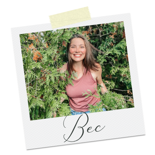 Smiling woman with long brown hair in a sleeveless pink top, surrounded by green foliage and branches.