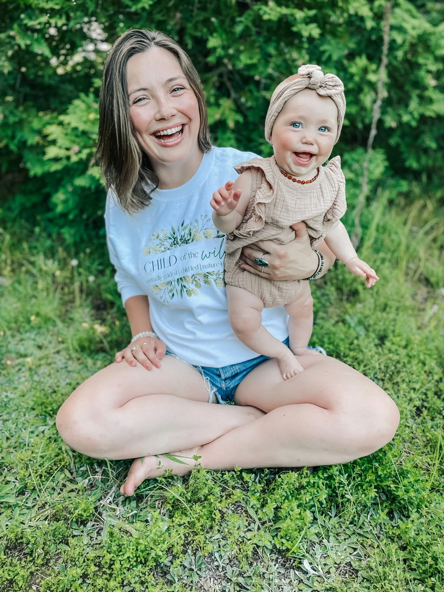 A woman and a baby girl sitting in a grassy outdoor area, smiling and enjoying the moment. The woman is sitting cross-legged and wearing a white t-shirt and shorts, while the baby girl is in a beige outfit with a matching headband and jewelry.