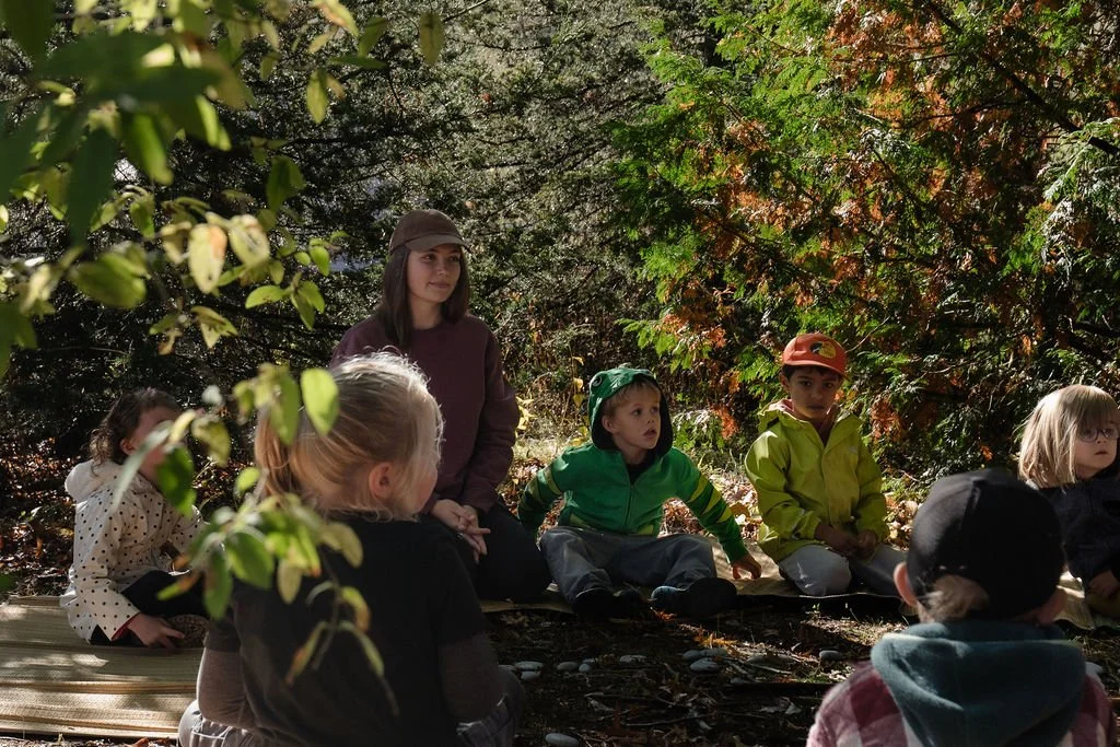 Children and a woman sitting outdoors on a wooden platform in a forested area during daytime.