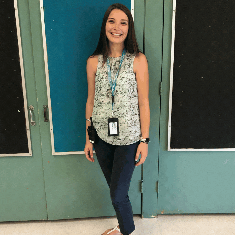 Young woman smiling, wearing a sleeveless patterned top, dark jeans, and flip-flops, standing in front of school lockers. She has a lanyard with ID badge and a walkie-talkie clipped to her waistband.