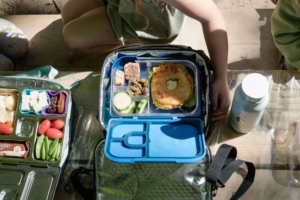 Child's school lunchbox with cucumber slices, pistachios, a bagel with cream cheese and a sliced apple, and a separate section with a bean, a square snack, and some dried fruit. A white bottle looks like a milk carton. The lunchbox is on a wooden table with other food containers nearby.