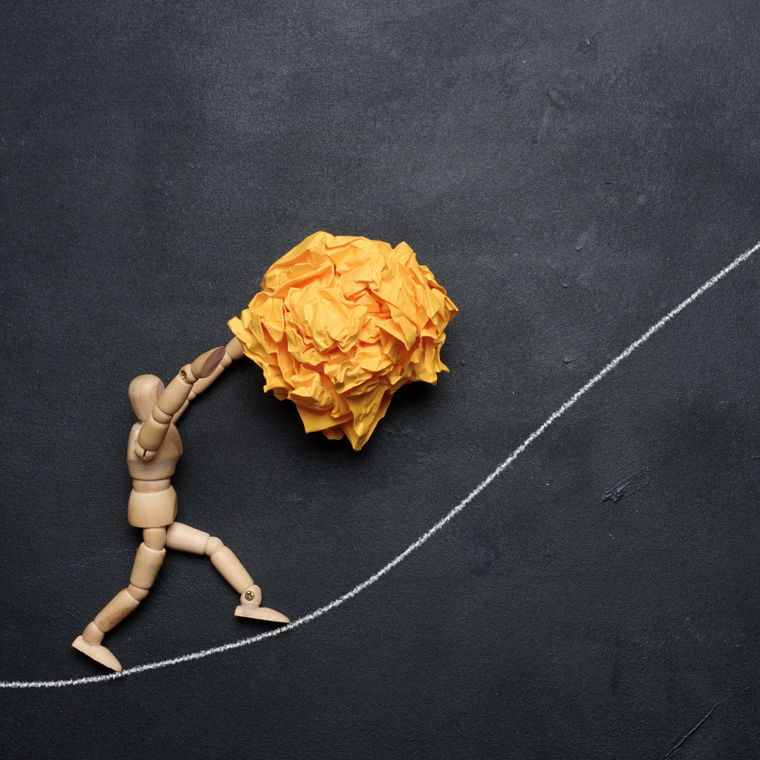 A wooden mannequin pushing a crumpled yellow paper ball up a white chalk-drawn upward sloping line on a black surface