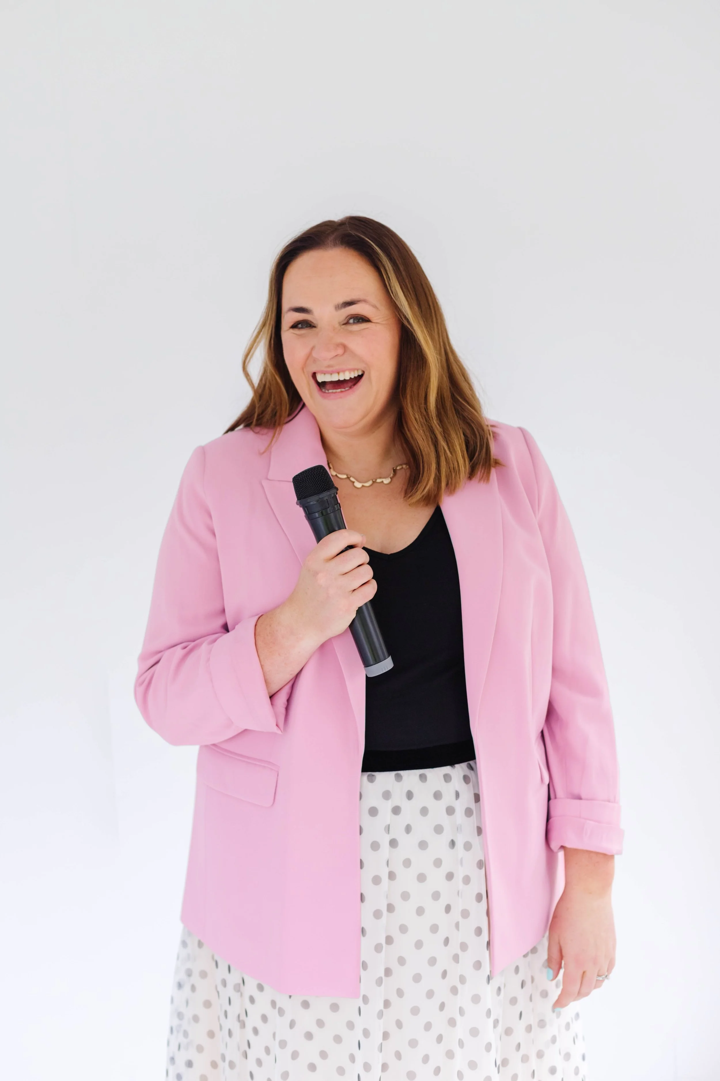 A motivational speaker wearing a pink blazer, black top, and polka dot skirt standing against a white wall, smiling and making a playful gesture. Engage your workforce to take control of their career. Leadership growth