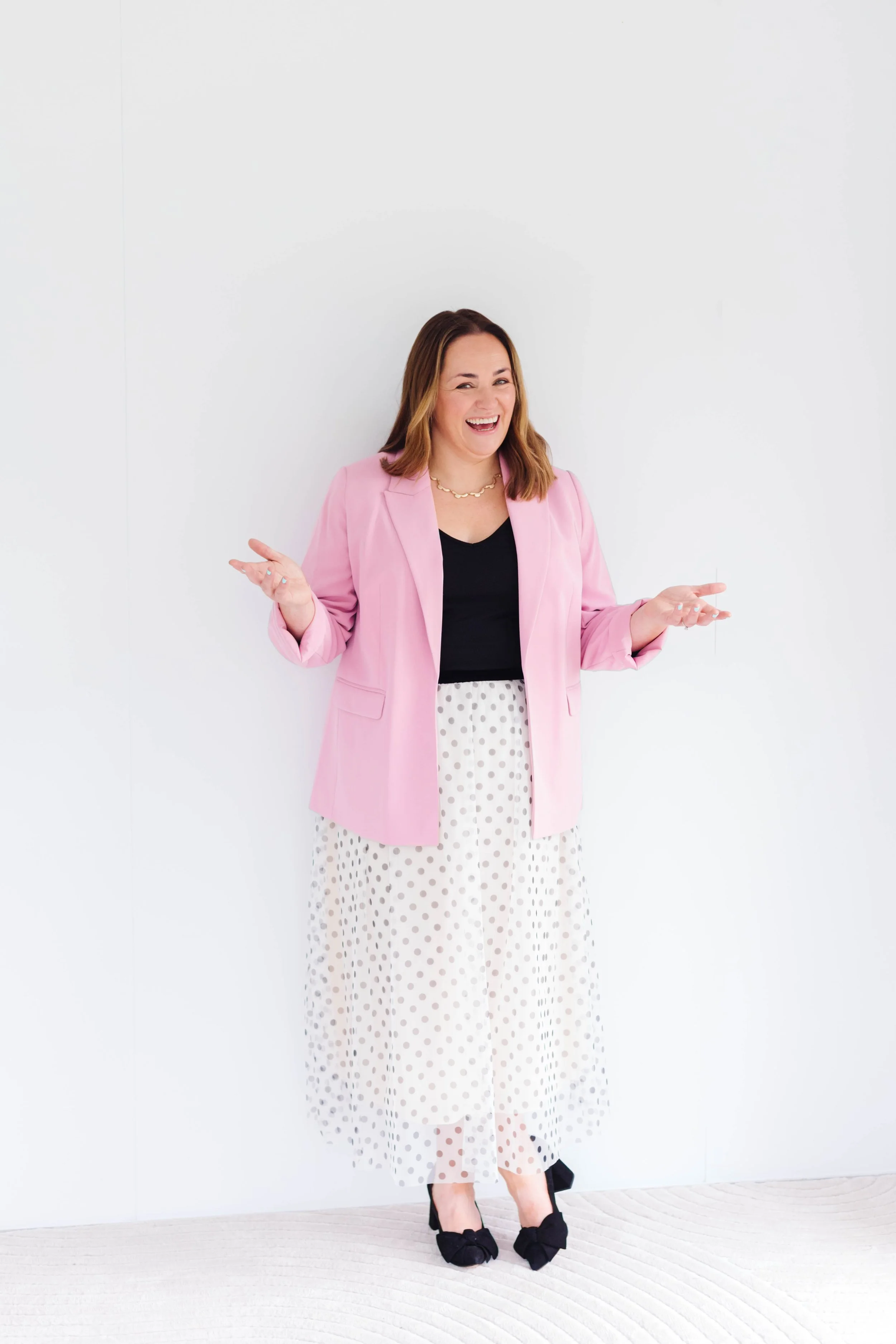 A leadership coach wearing a pink blazer, black top, and polka dot skirt standing against a white wall, smiling and making a playful gesture. 1 1 coaching for career challenges and finding balance in life. Midlife crisis tools.