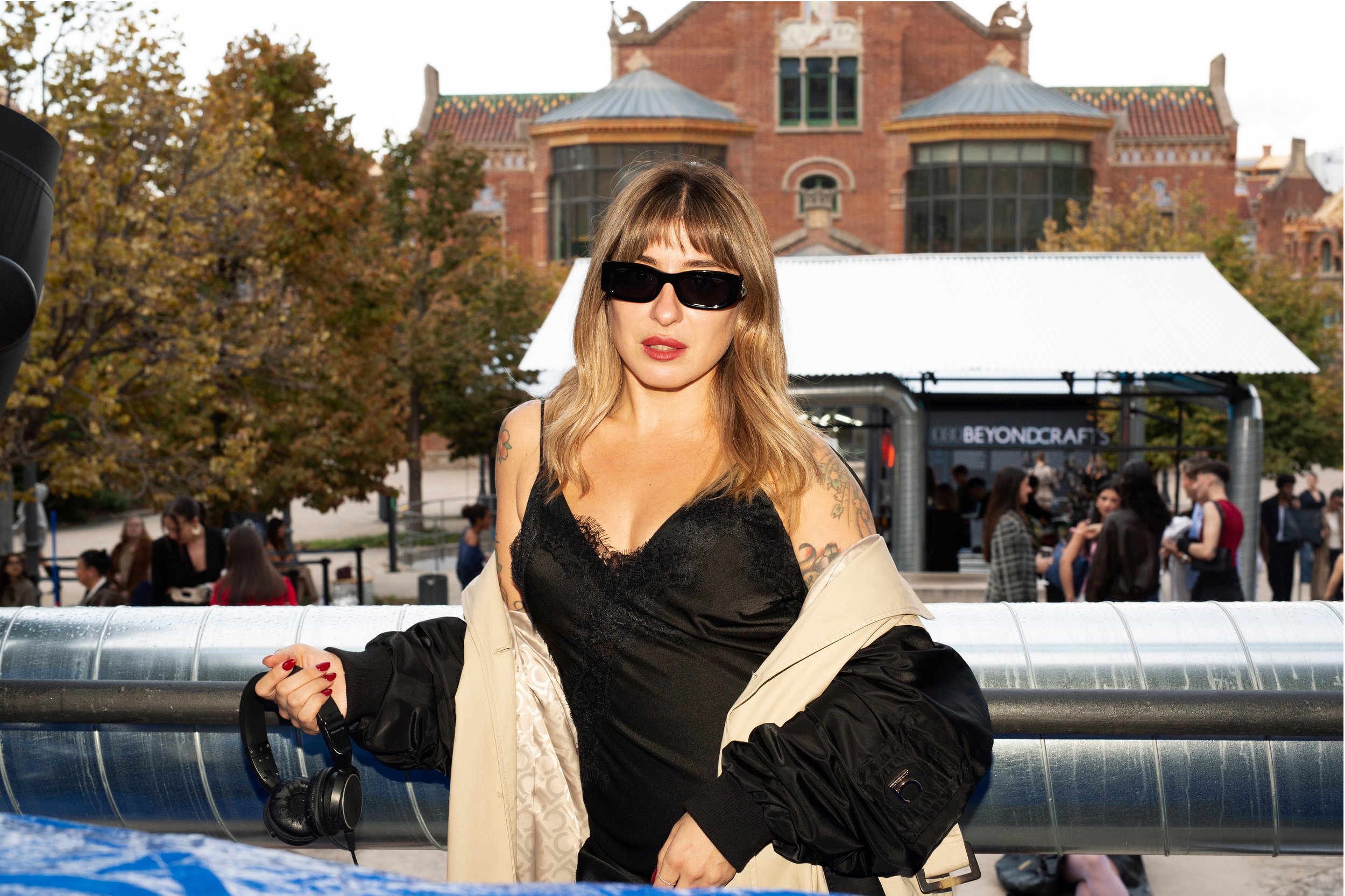 Mujer joven con gafas de sol, cabello rubio y vestido negro, sosteniendo audífonos, en un espacio al aire libre con edificios y árboles en el fondo.