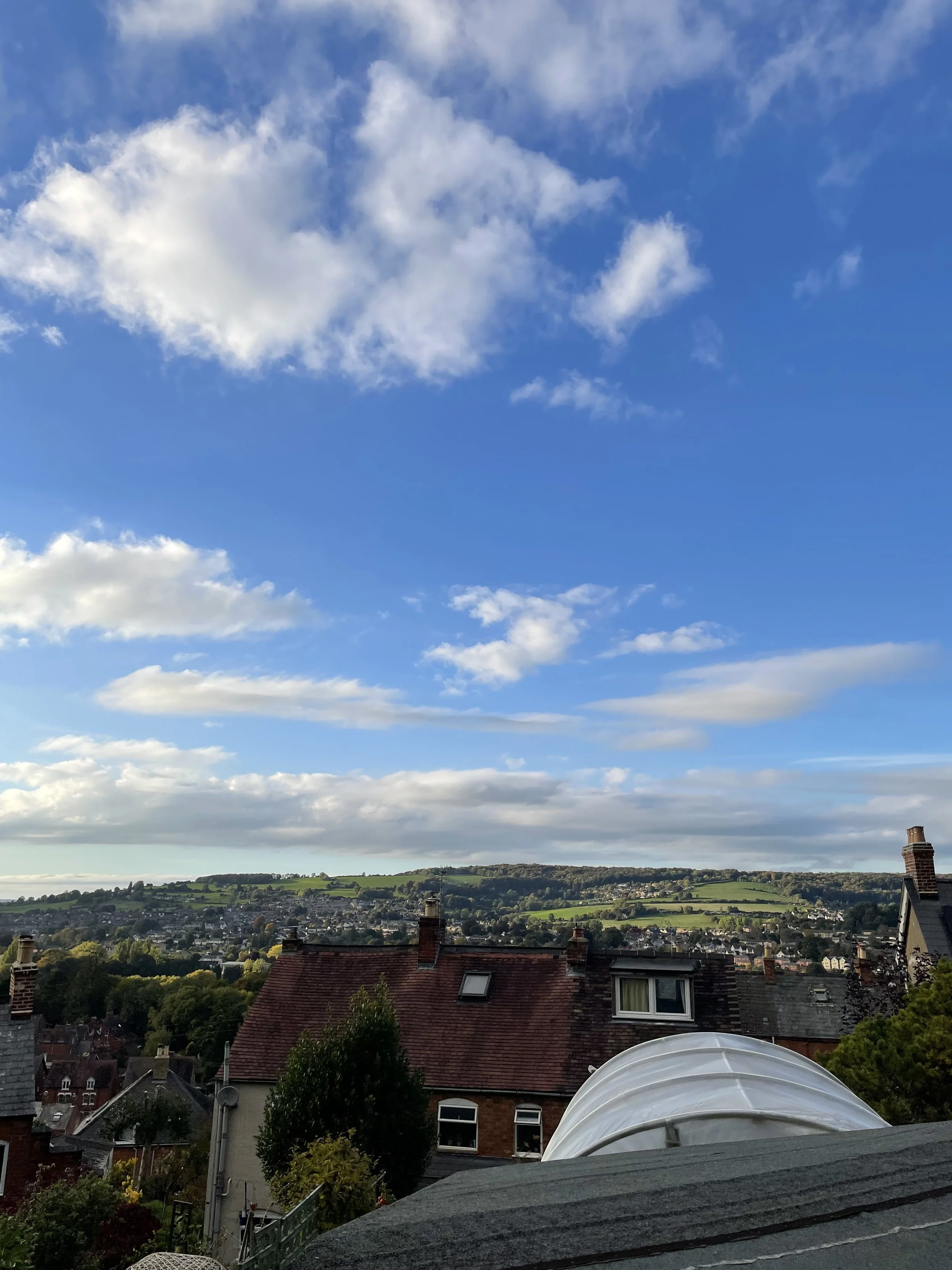 View of a residential neighborhood with rooftops, trees, and a distant hillside under a partly cloudy blue sky.