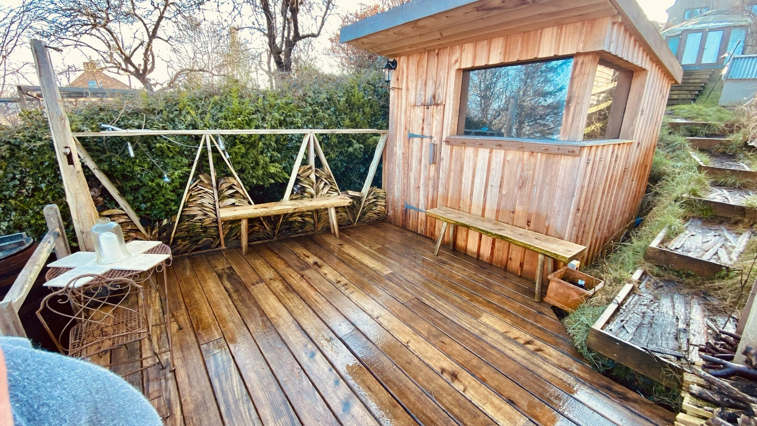 Wooden deck with a wooden sauna, a bench, firewood stacked against a hedge and a hillside with stairs.