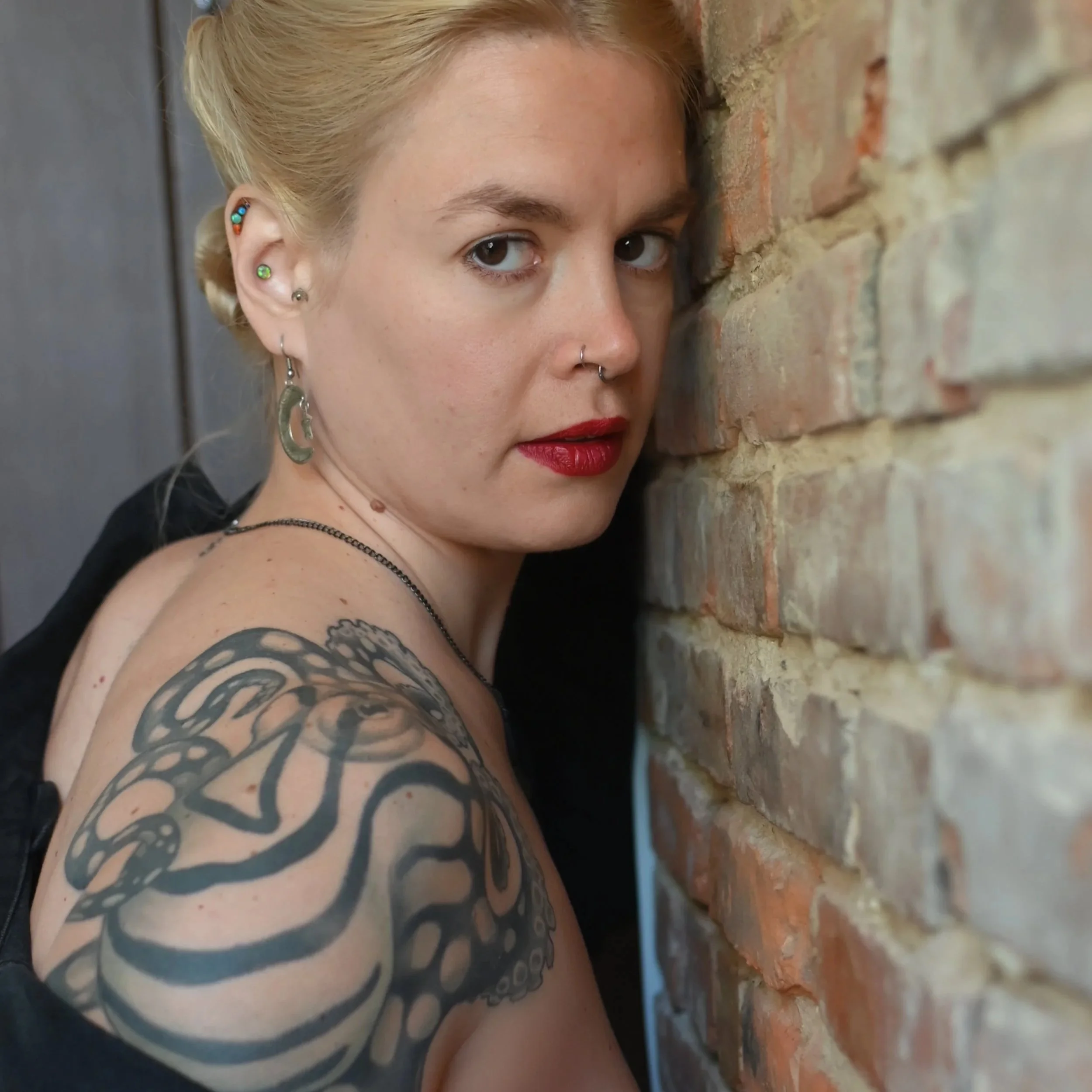 Close-up of a Goddess Hecate with blonde hair, multiple earrings, red lipstick, and a large black octopus tattoo on her shoulder, standing against a brick wall.