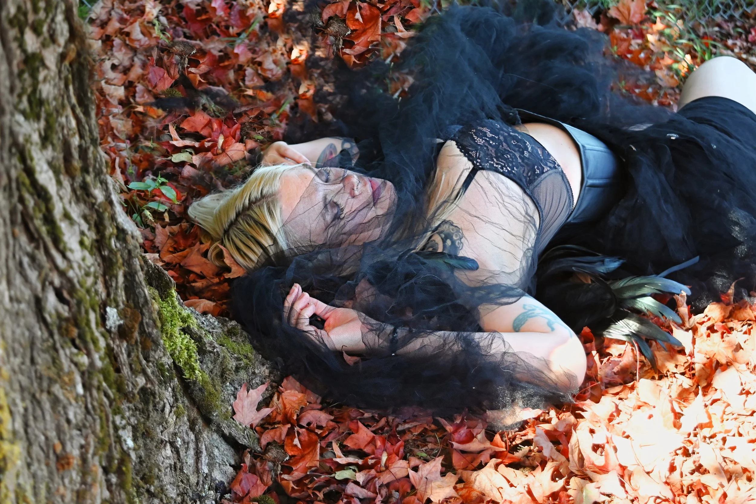 A woman lying on her side on the forest ground covered with autumn leaves, wearing a black lace top, black tulle skirt, and thigh-high stockings, with tattoos on her arm and chest, and blond hair. She is touching her head with her hand and appears to be relaxed or sleeping in a natural outdoor setting.