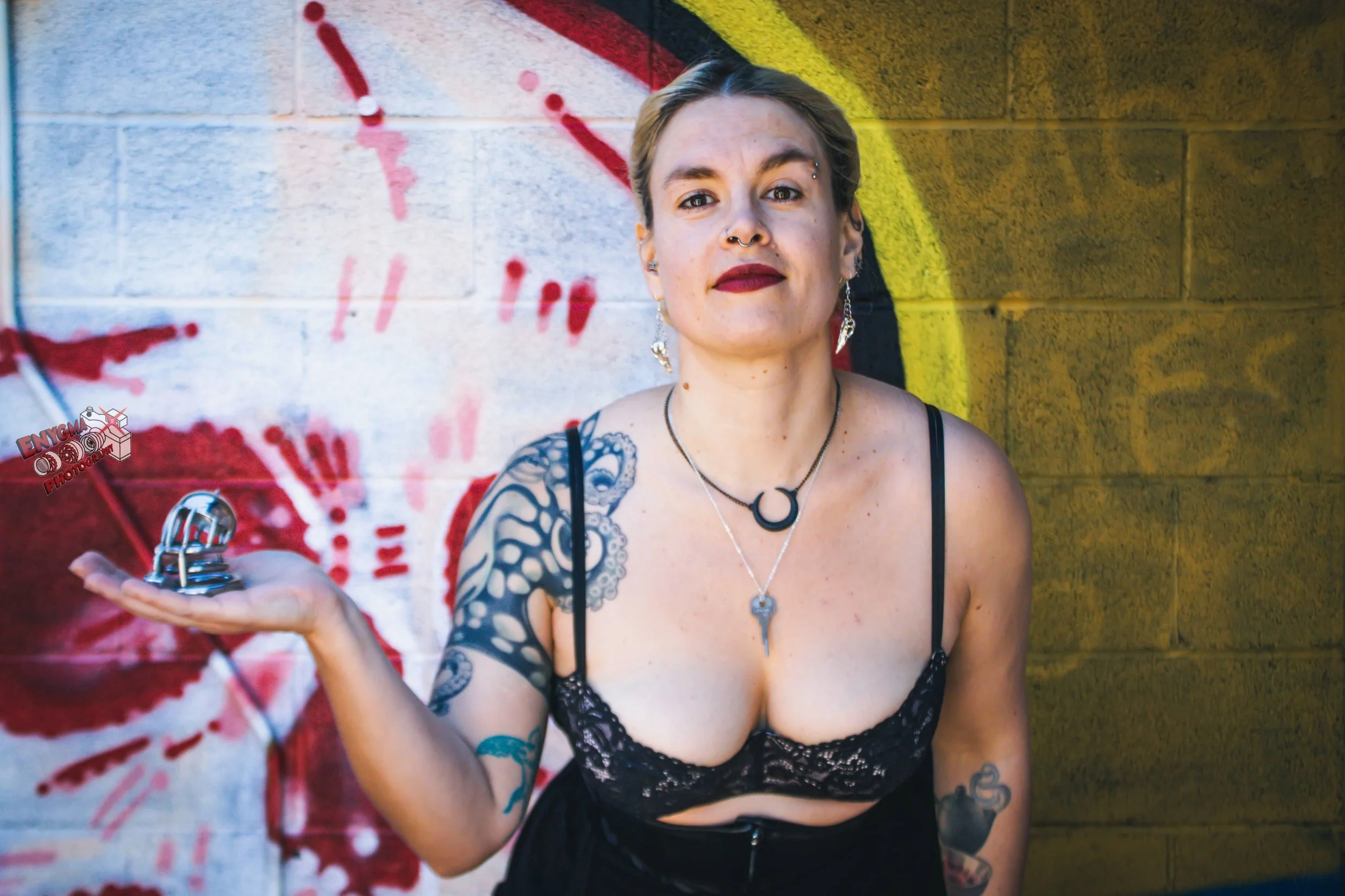 Goddess Hecate, a professional Dominatrix stands in front of a graffiti wall, holding a small metallic object in her outstretched hand. It is a male chastity cage. She is wearing a black lace top and jewelry, with colorful background street art.