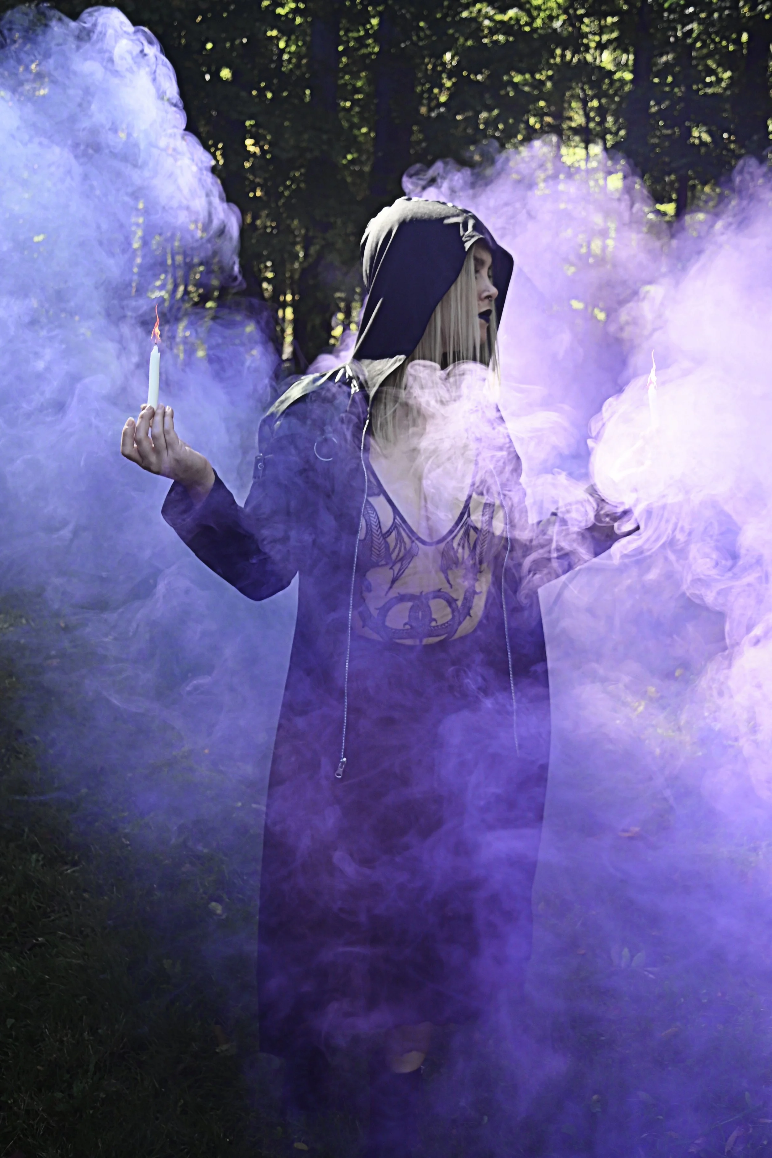 A witch and professional Dominatrix named Goddess Hecate dressed in dark clothing and a hood, holding lit candles, standing amidst purple smoke in an outdoor wooded area during the day.