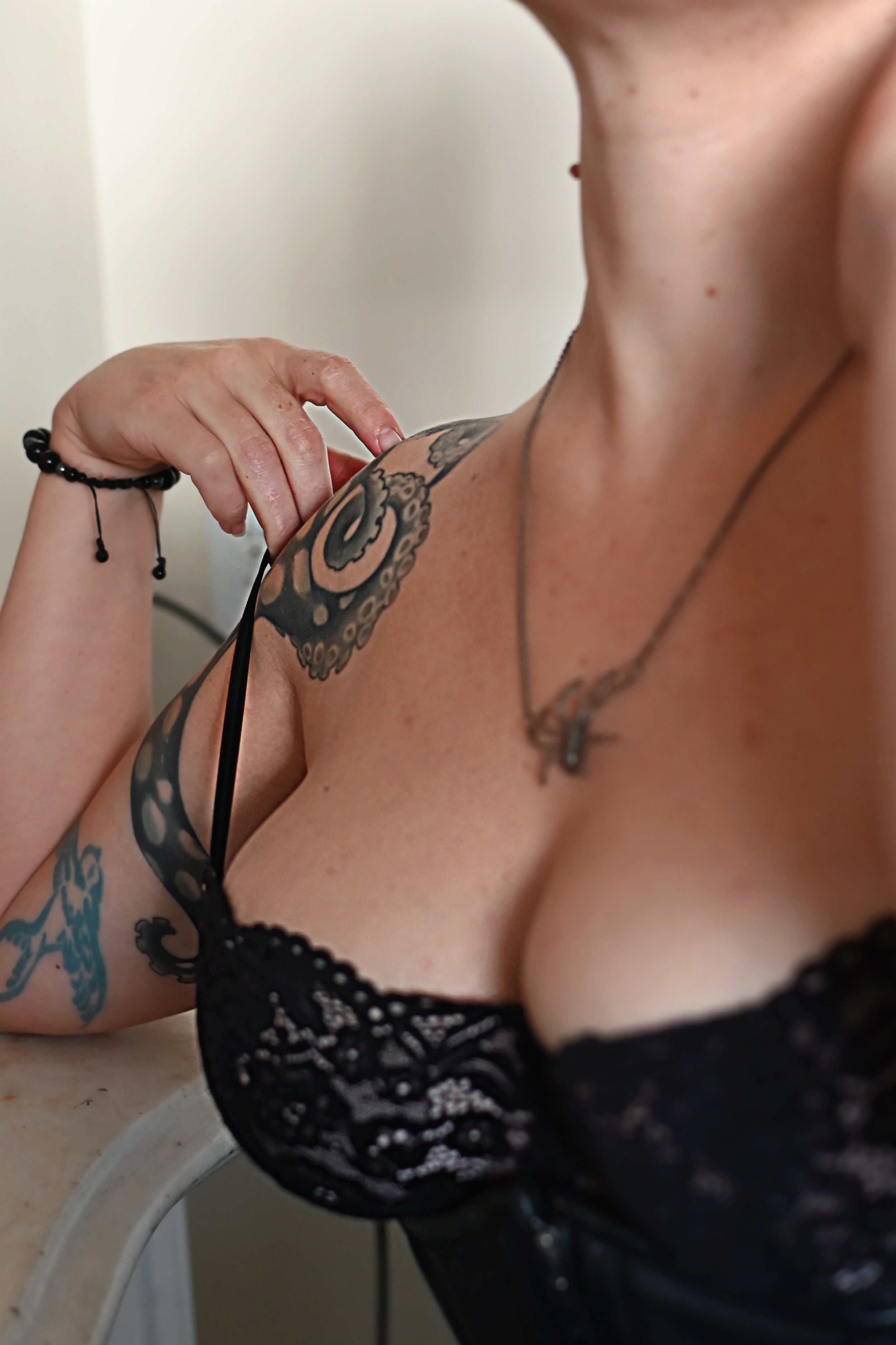Close-up of a woman with tattoos, wearing a black lace top, holding the strap of her top, with a necklace and bracelet visible.