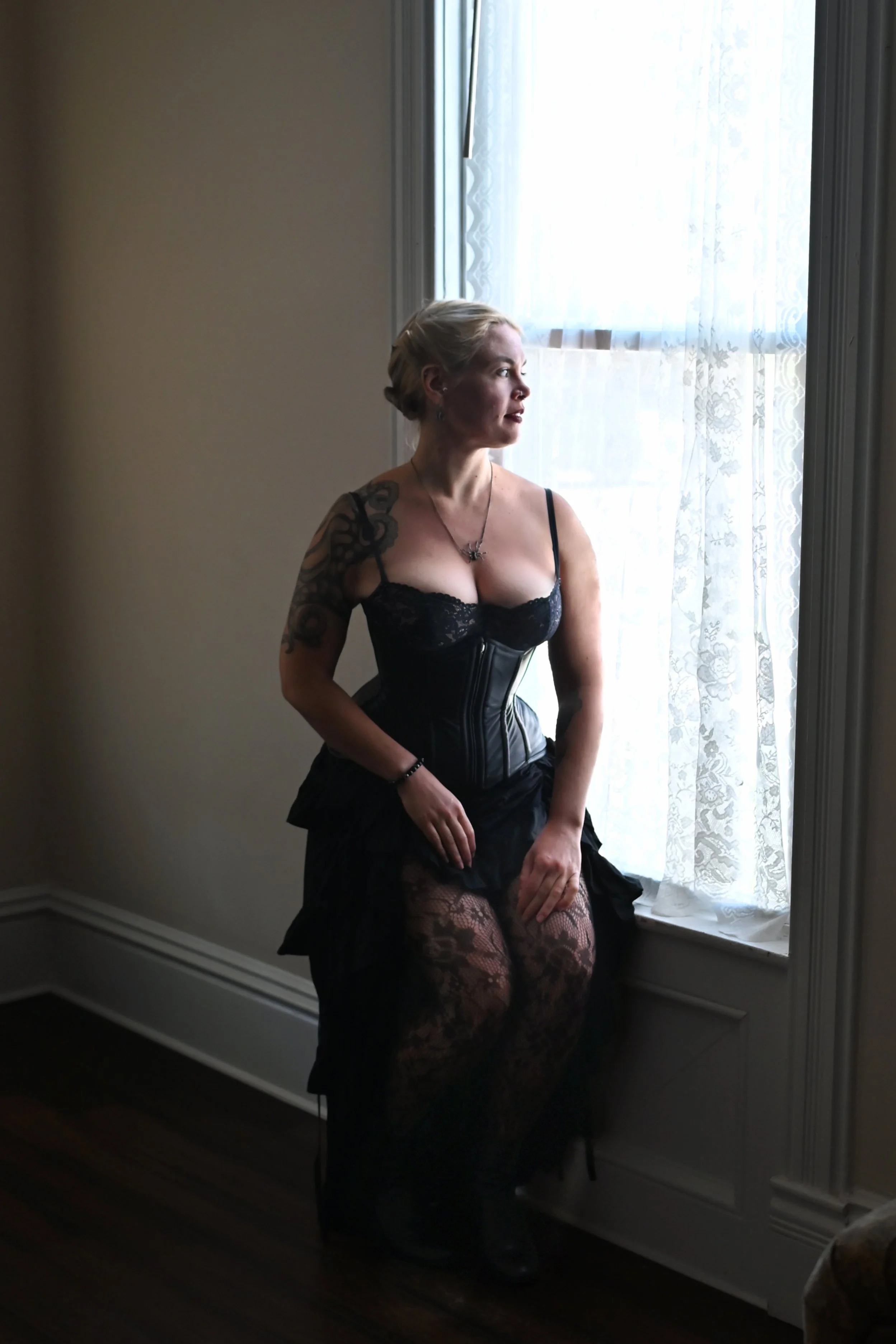 A woman sitting on a windowsill in lace tights, a black lace top, and a leather corset, looking out the window with soft lighting.