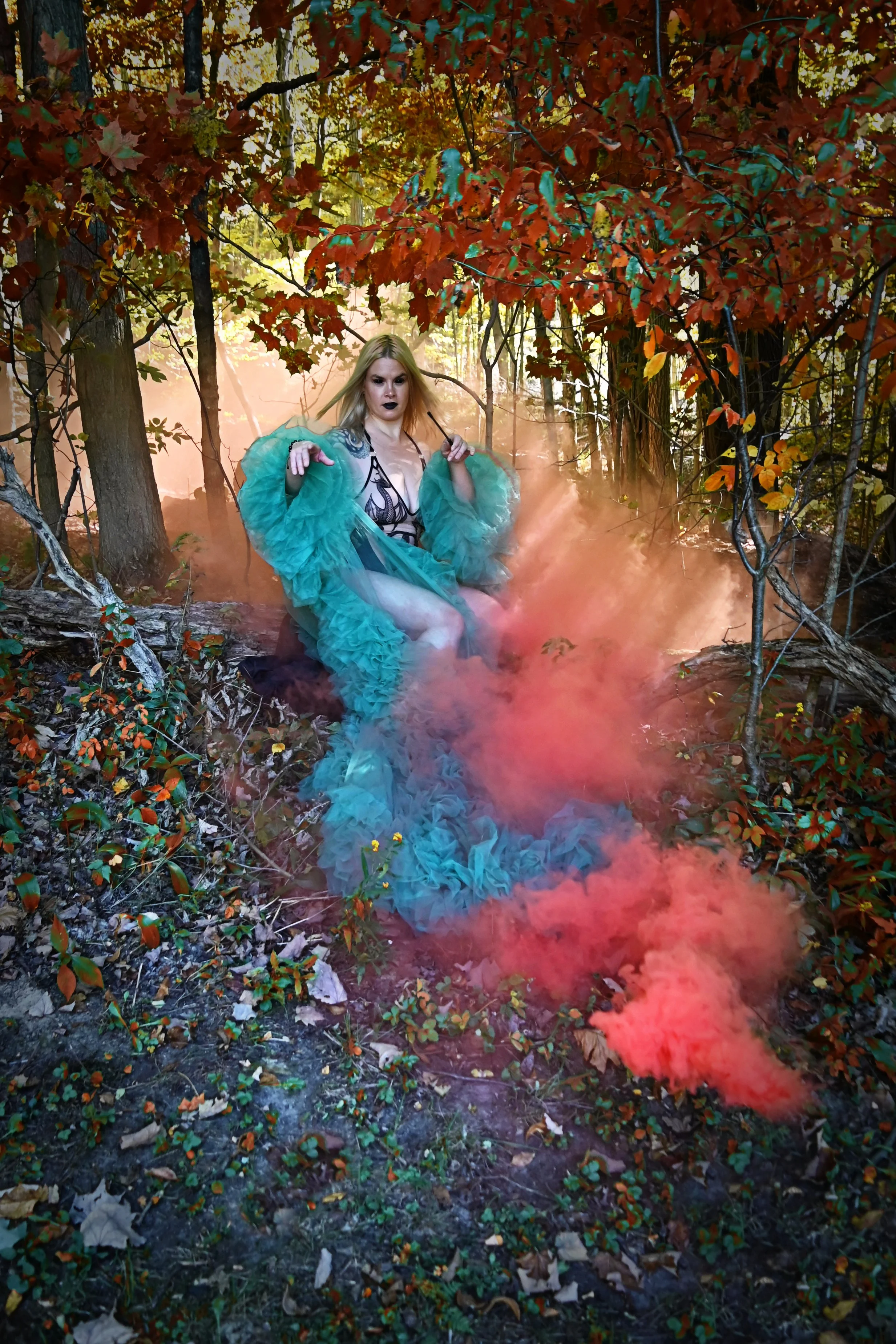 A powerful witch and pro Domme named Goddess Hecate in a teal ruffled dress and black lingerie top sitting in a forest surrounded by orange autumn leaves, with orange smoke around her.