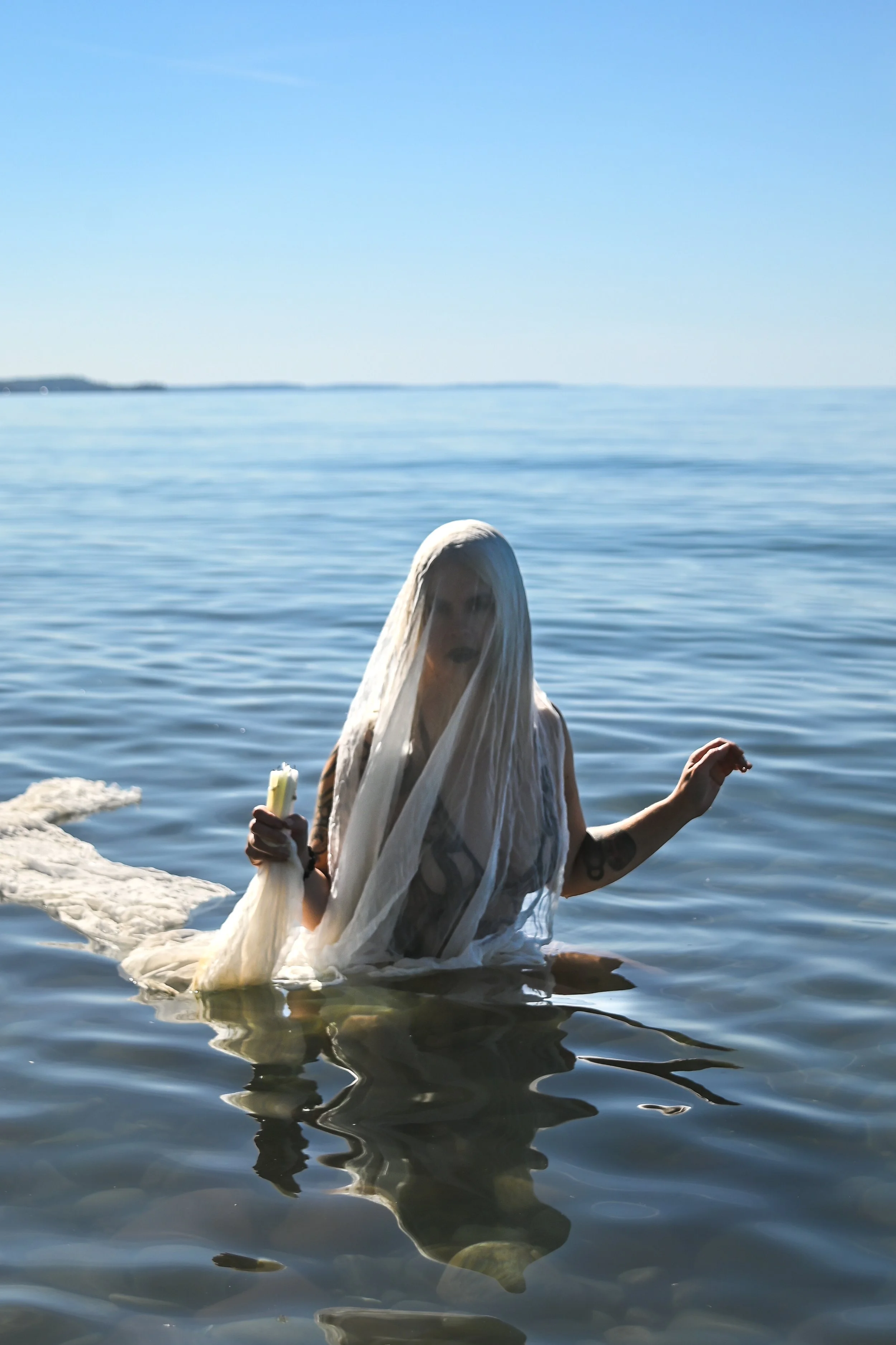A woman with long white hair wearing a white veil and white clothing emerges from ocean water holding a candle, with a serene ocean and blue sky background.