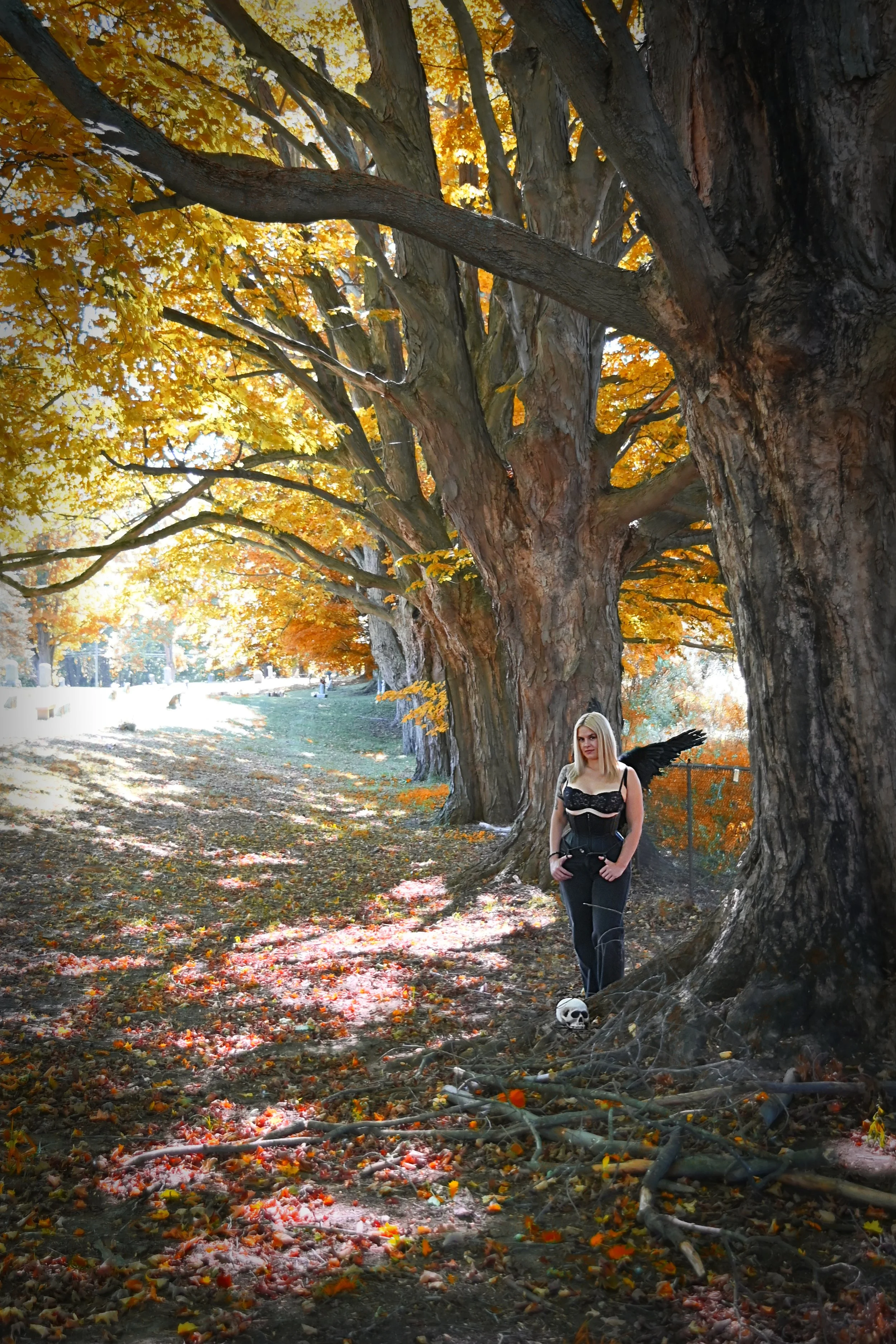 Female Dominatrix Goddess Hecate dressed as a dark angel with black wings, short blond hair, and gothic attire standing in a cemetery next to a large tree with an orange and yellow autumn canopy in a park.